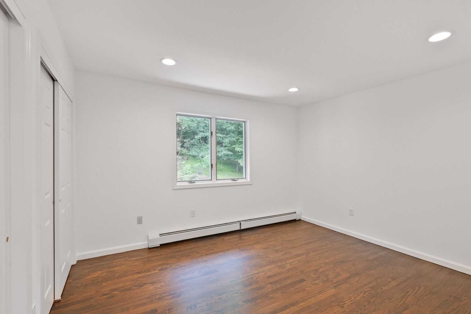 Empty room with hardwood floors, white walls, closet, window, and recessed lighting.