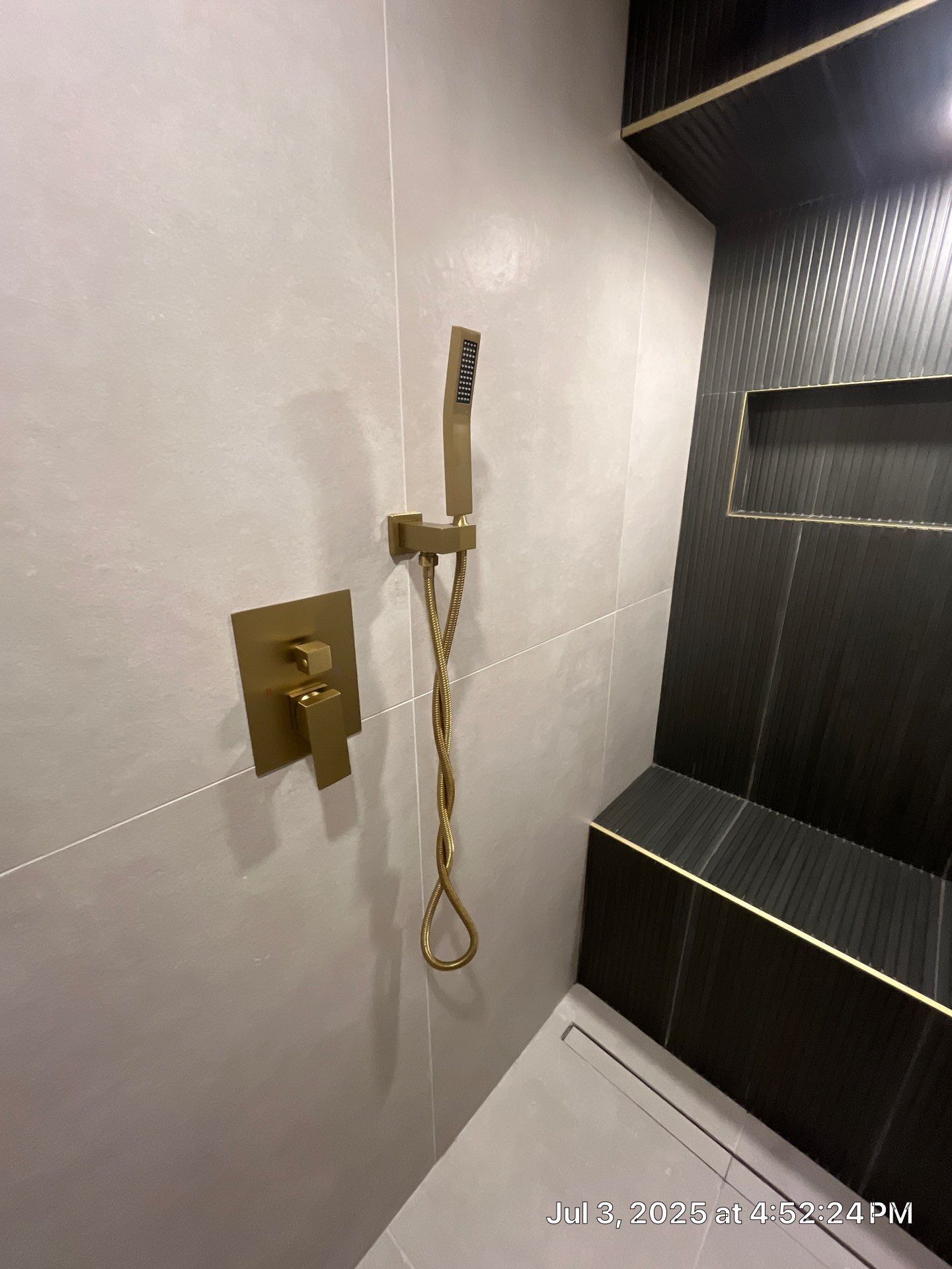 Gold shower fixtures on light gray tiled wall; black tiled alcove with gold trim.