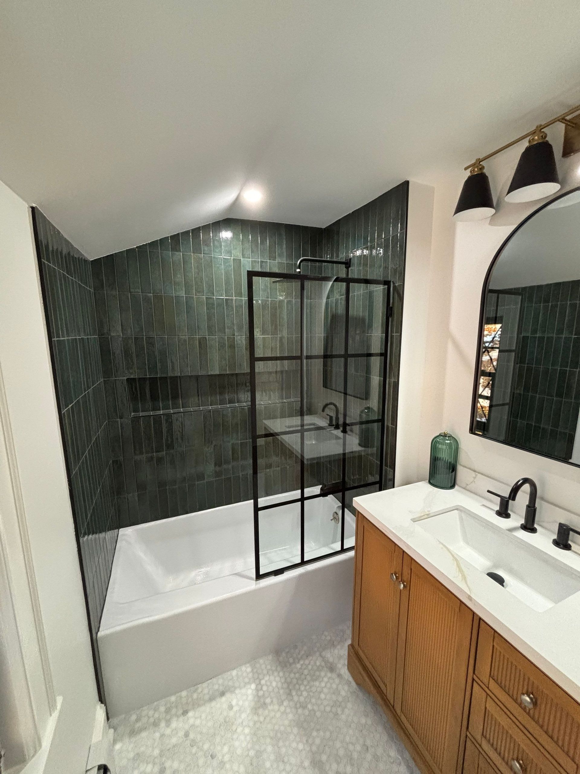 Bathroom with green tile shower, black framed glass door, white sink and vanity, and brass light fixtures.