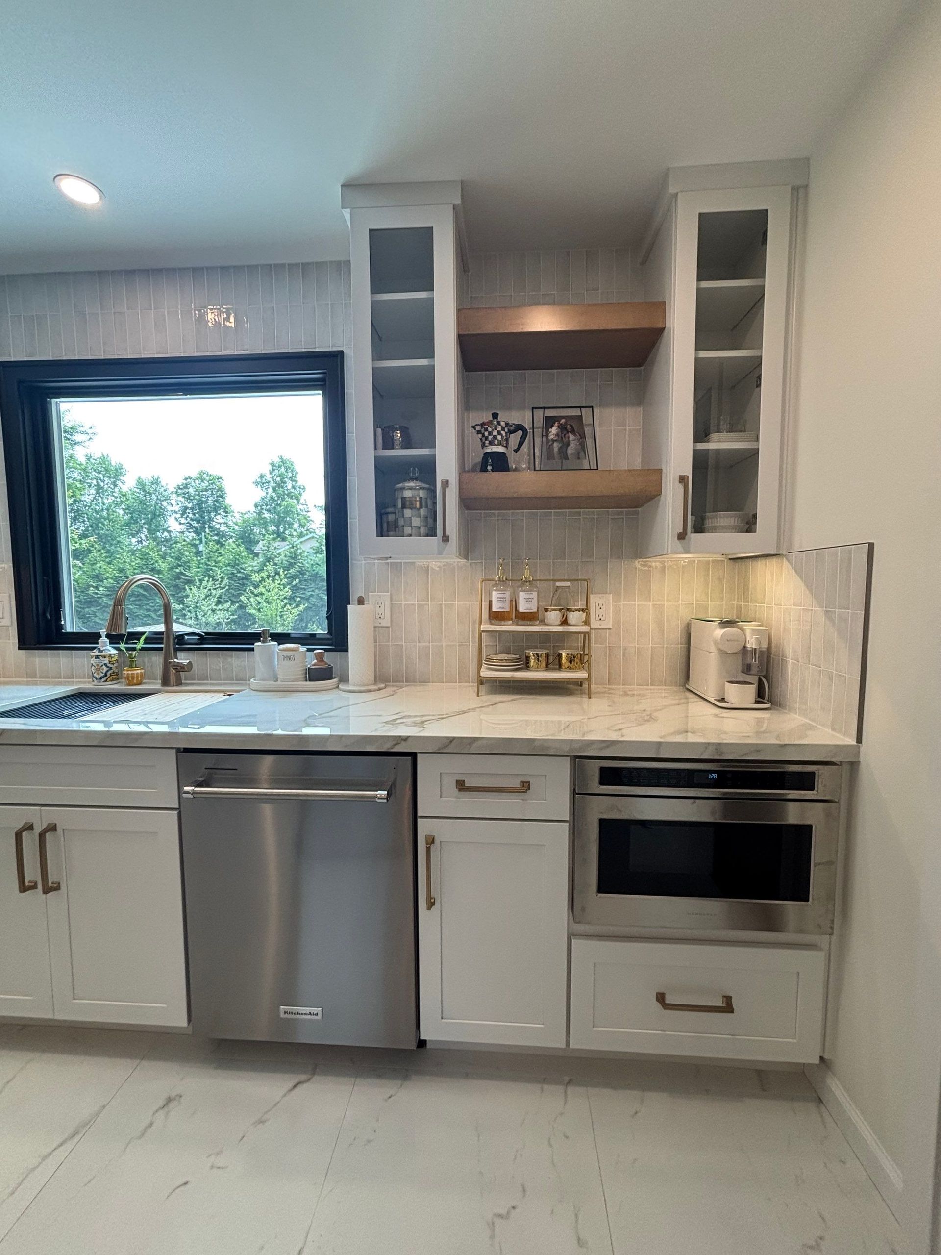 Modern white kitchen with stainless steel appliances, marble countertops, and a window overlooking greenery.