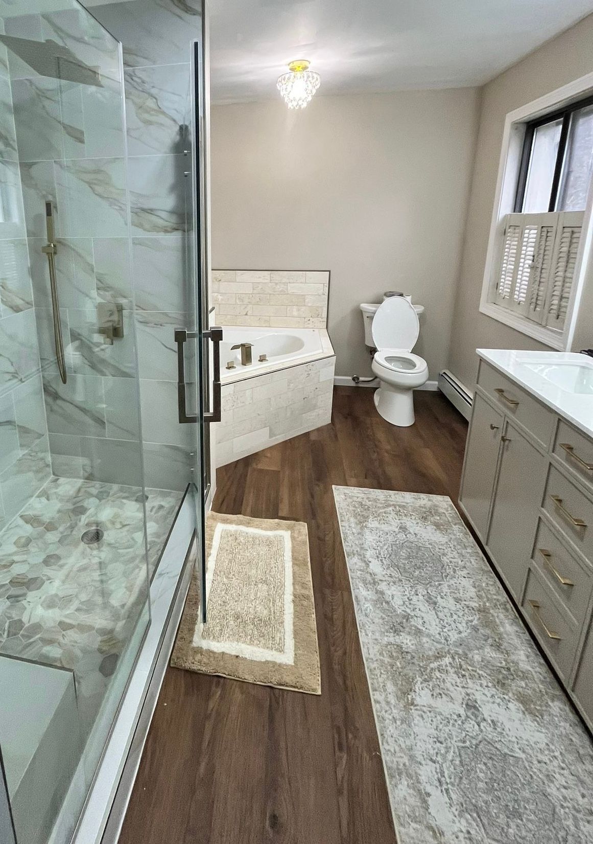 Bathroom with a glass shower, tub, toilet, vanity, and rugs on wood-look flooring.