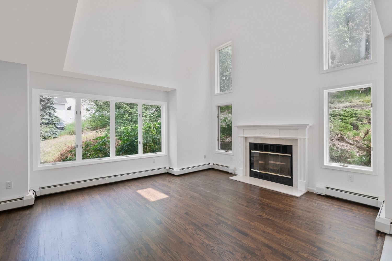 Bright, empty living room with large windows overlooking trees, fireplace, and dark wood floors.