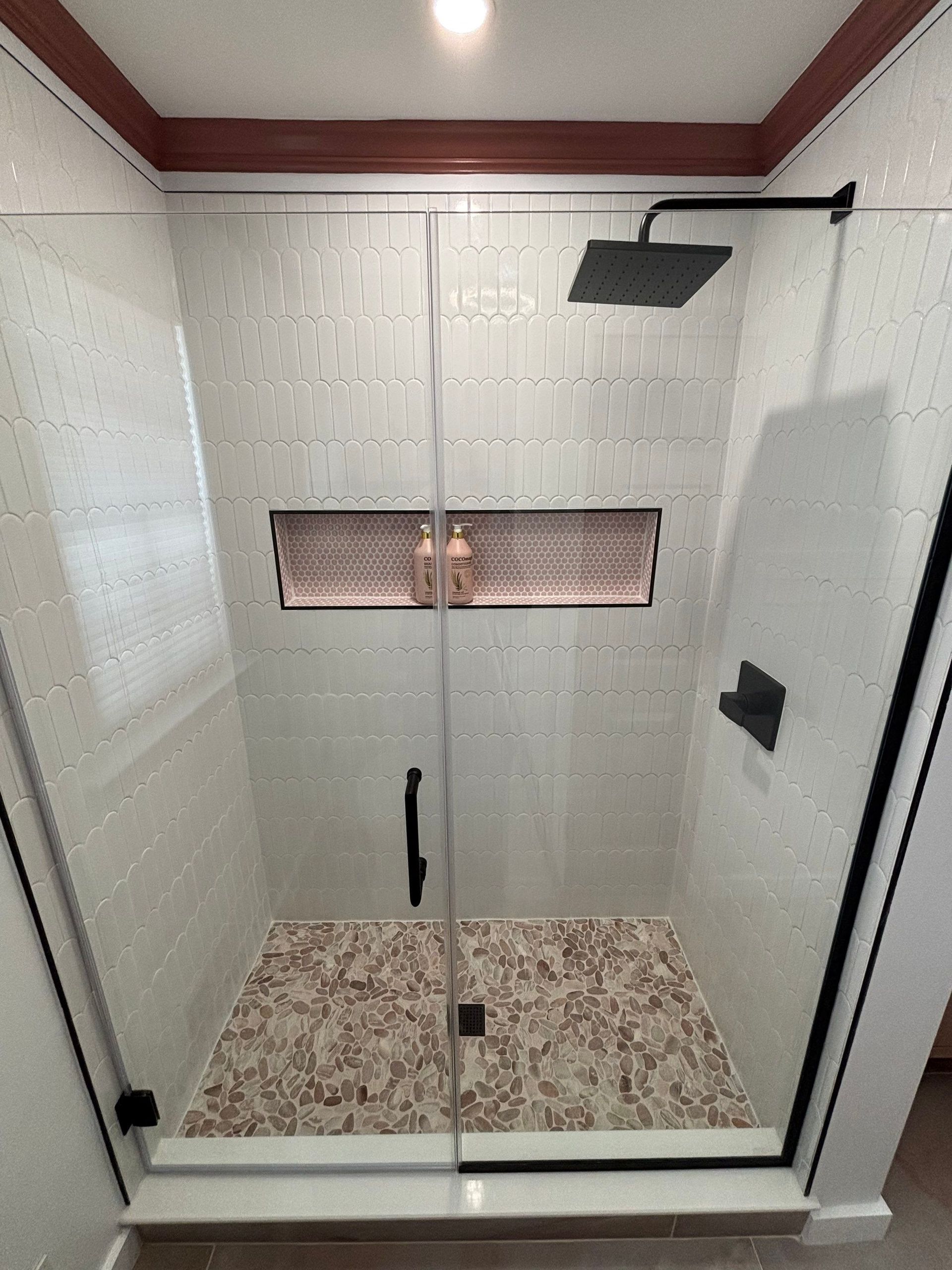 Shower with white patterned tiles, glass door, black fixtures, and stone floor.