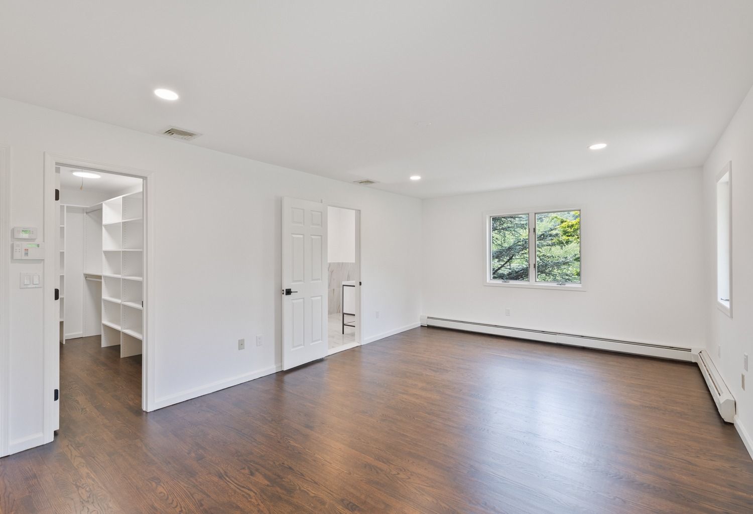 Empty bedroom with hardwood floors, white walls, closet, and a window overlooking greenery.