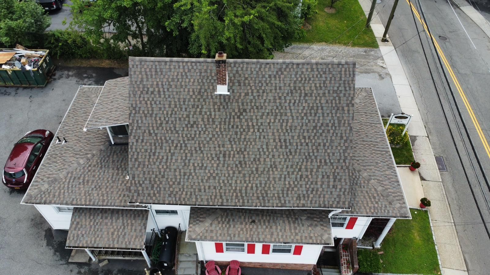 Overhead view of a building with a gray shingled roof, chimney, and red shutters, next to a street.