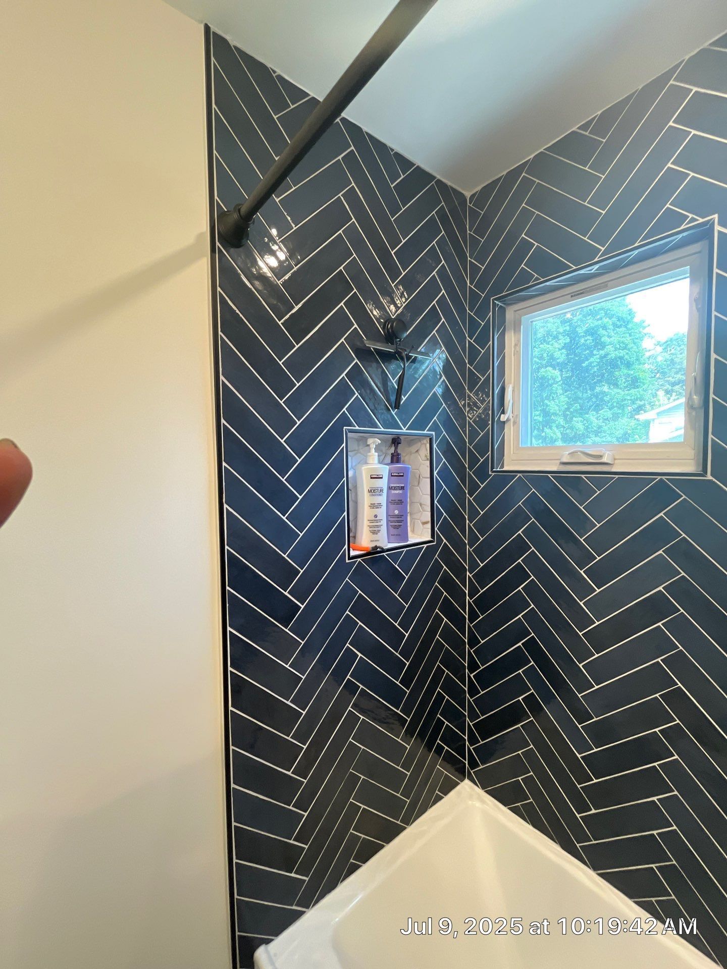 Blue herringbone tiled shower, small window, dark showerhead and soap dispenser.