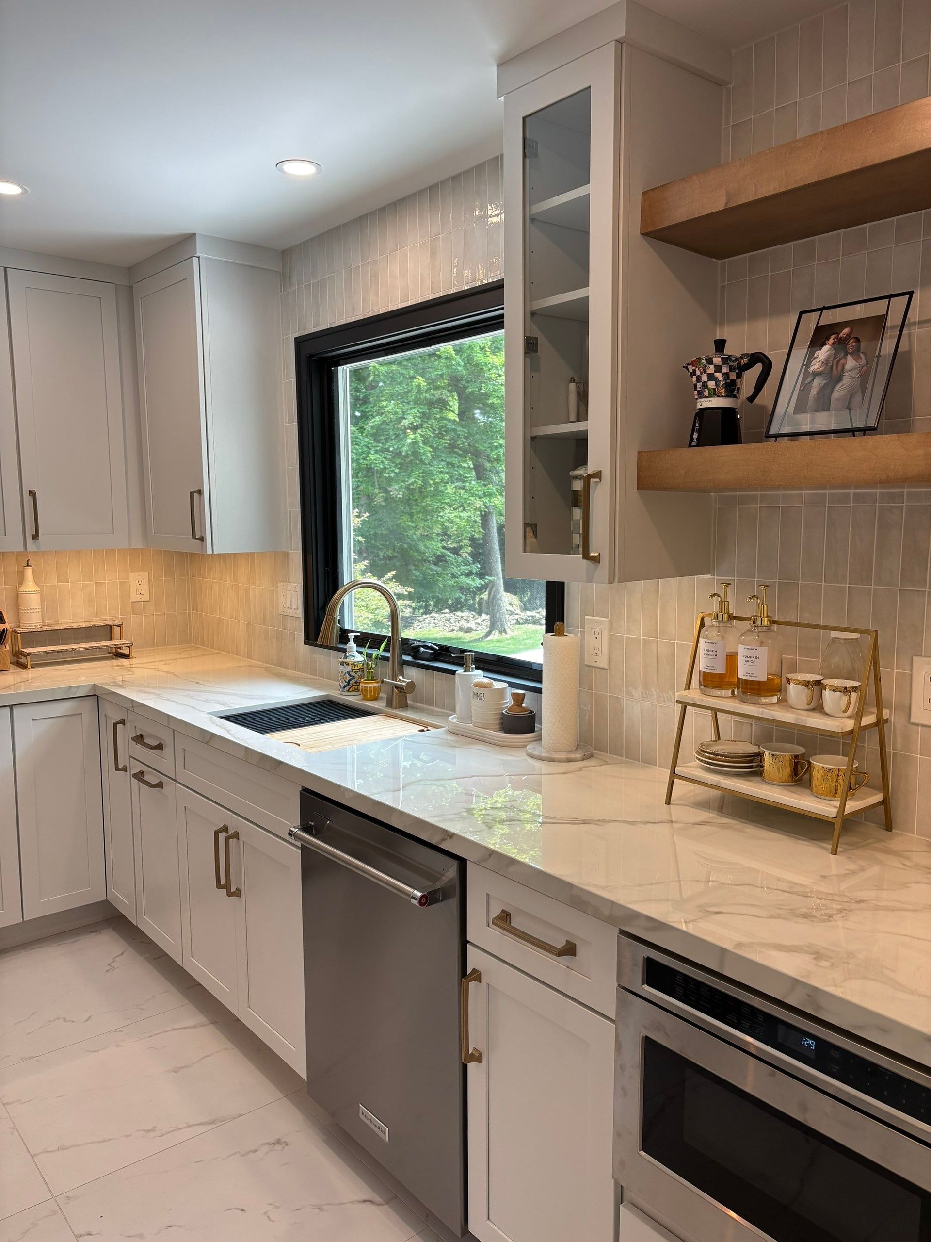 Modern white kitchen with stainless steel appliances, marble countertops, and a window.