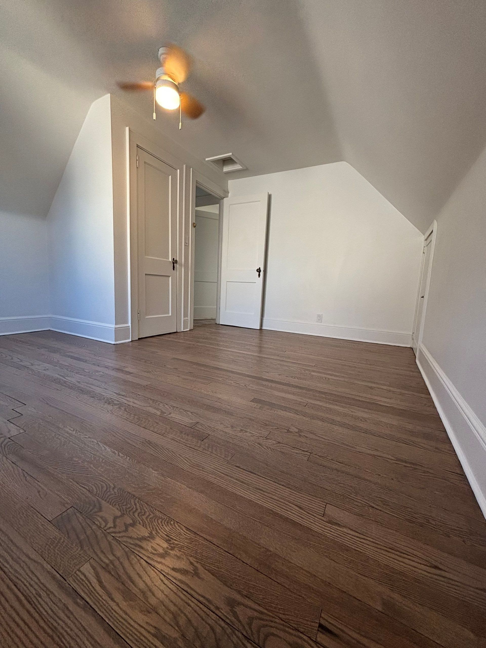 Empty attic bedroom with hardwood floor, white walls, two doors, and a ceiling fan.