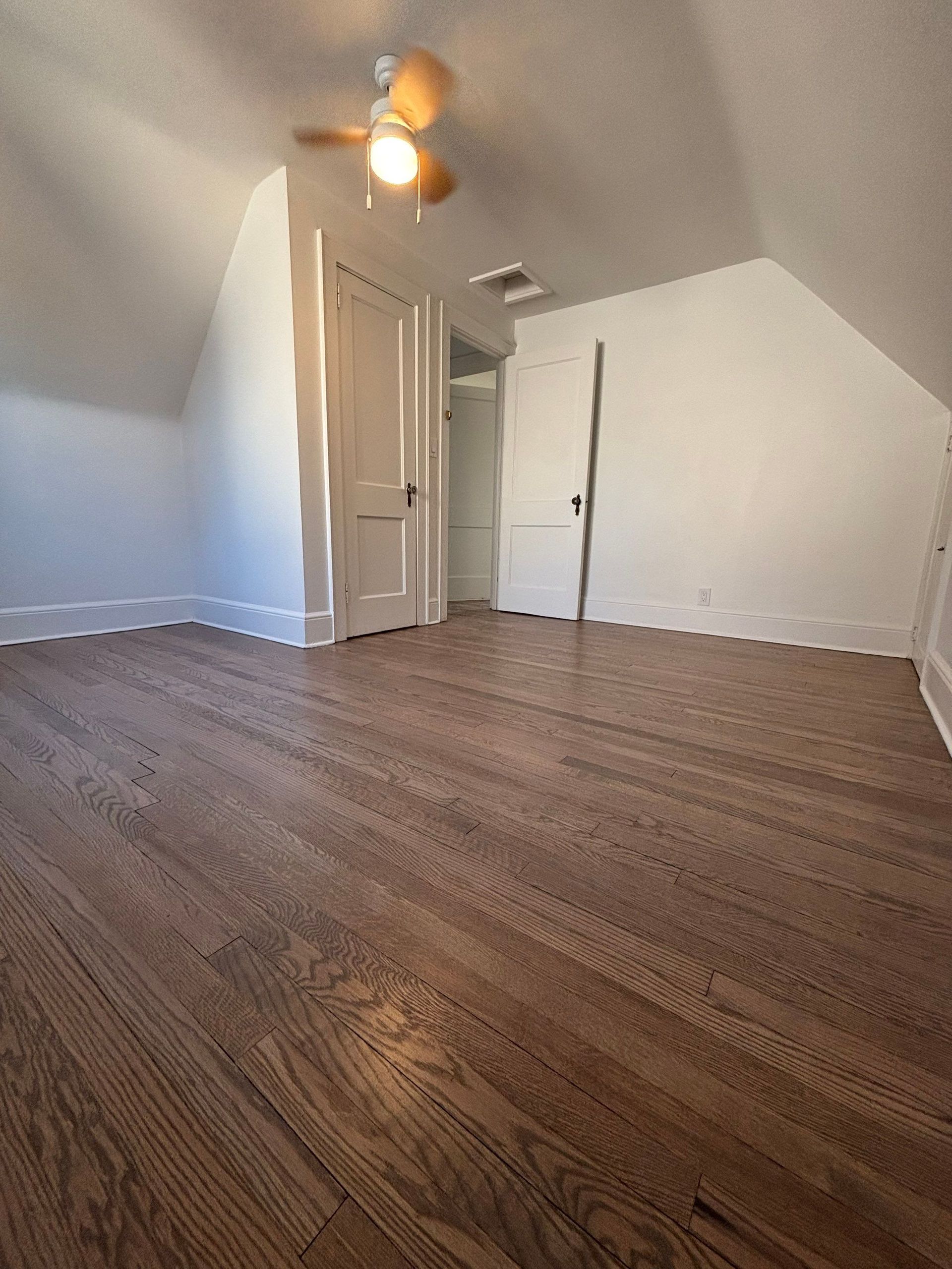 Empty attic bedroom with wood floor, white walls and doors, a ceiling fan.