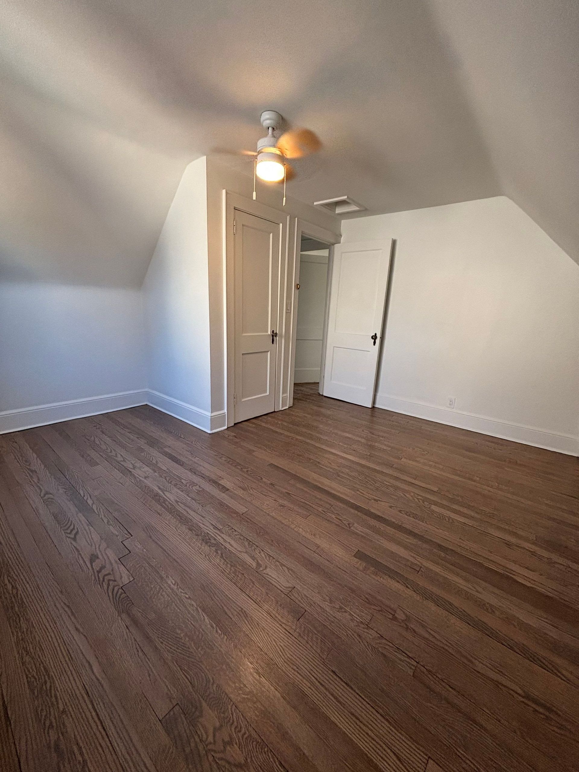 Empty room with hardwood floors, white walls, and a sloped ceiling. A closed door and a doorway are visible.
