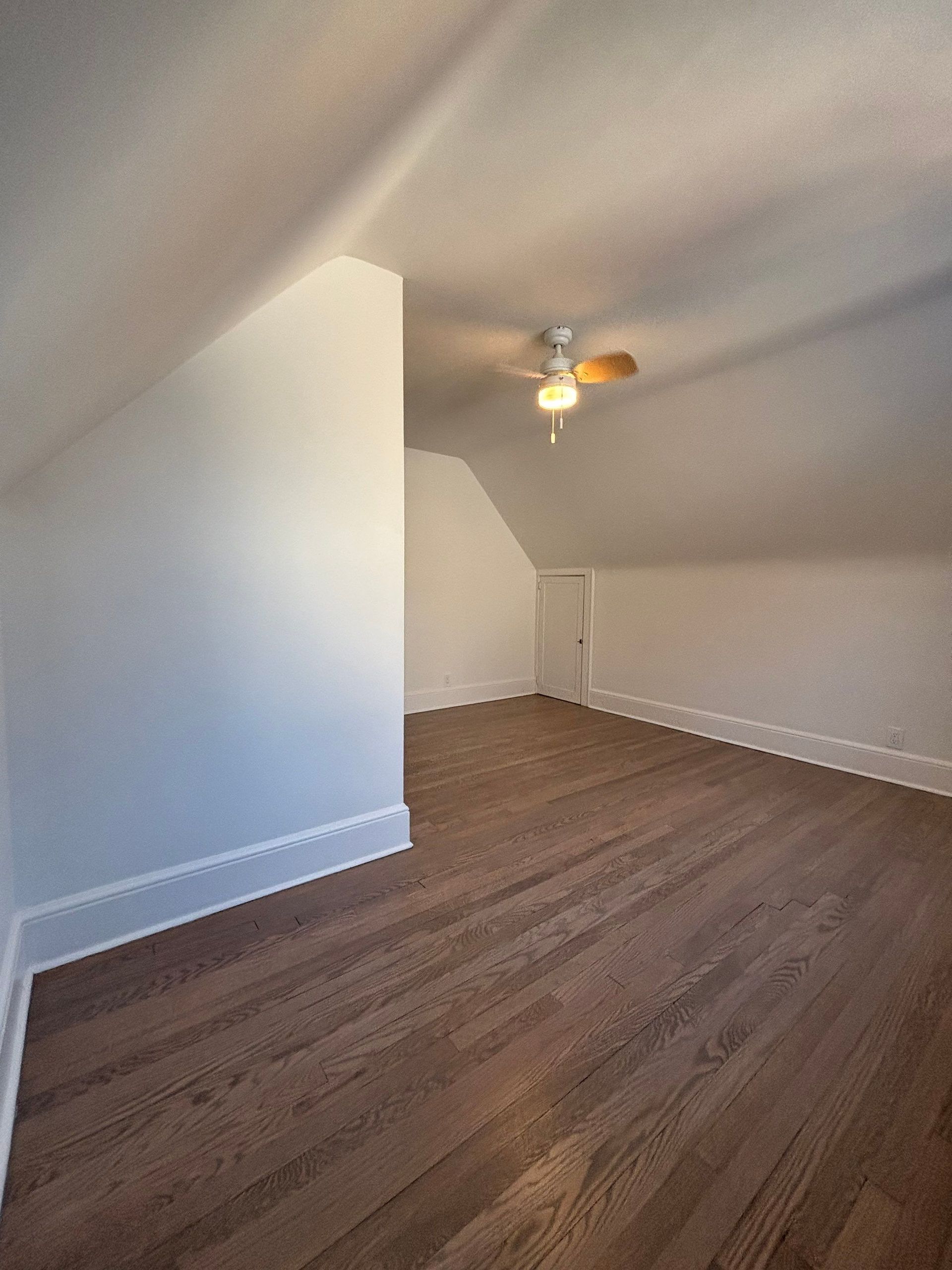 Empty attic room with wood floor and sloped walls. Ceiling fan in center.