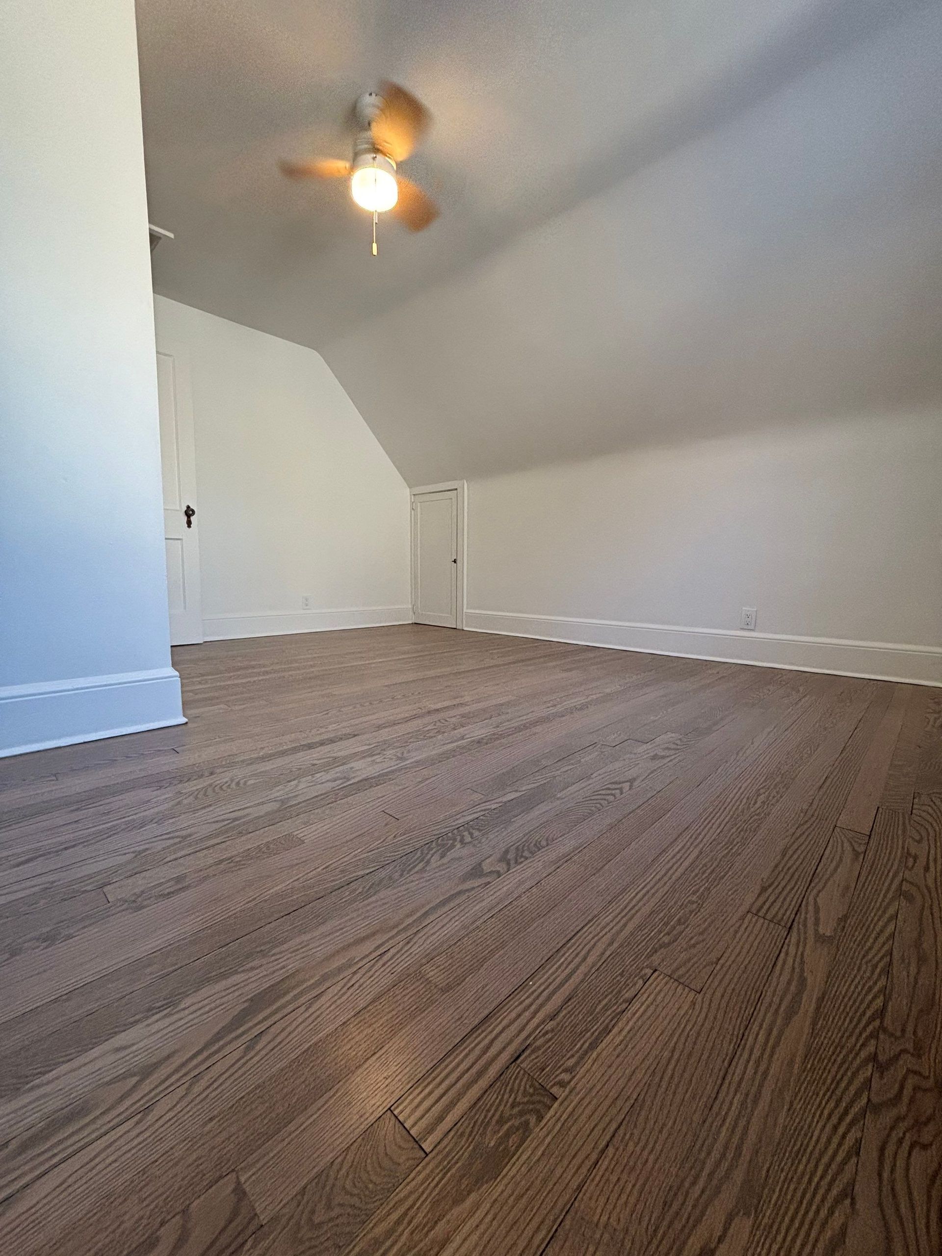 Empty attic room with brown wood-look flooring, sloped ceiling, and a ceiling fan.
