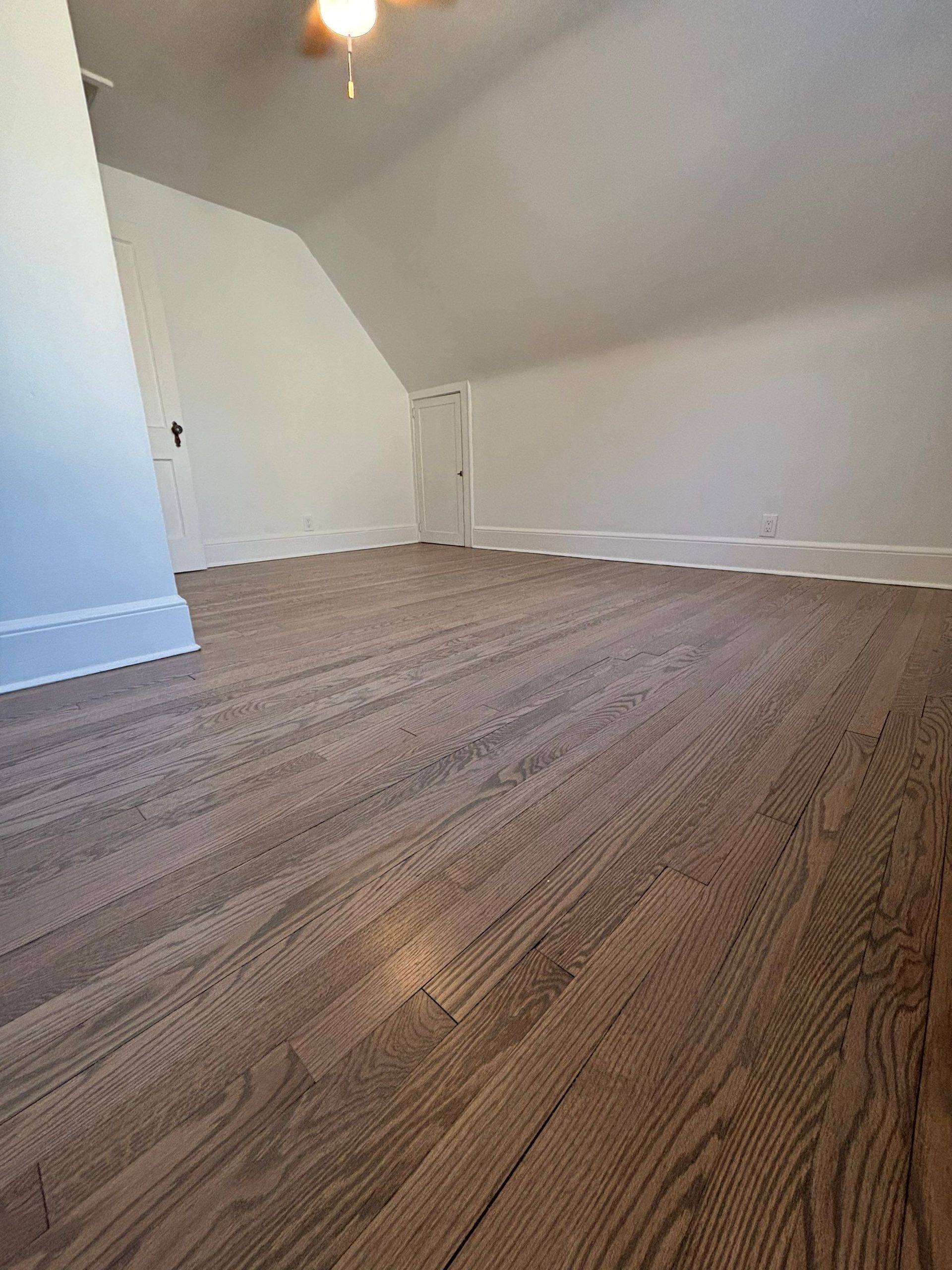 Room with angled ceiling, wood-look flooring, white walls, and a small door.