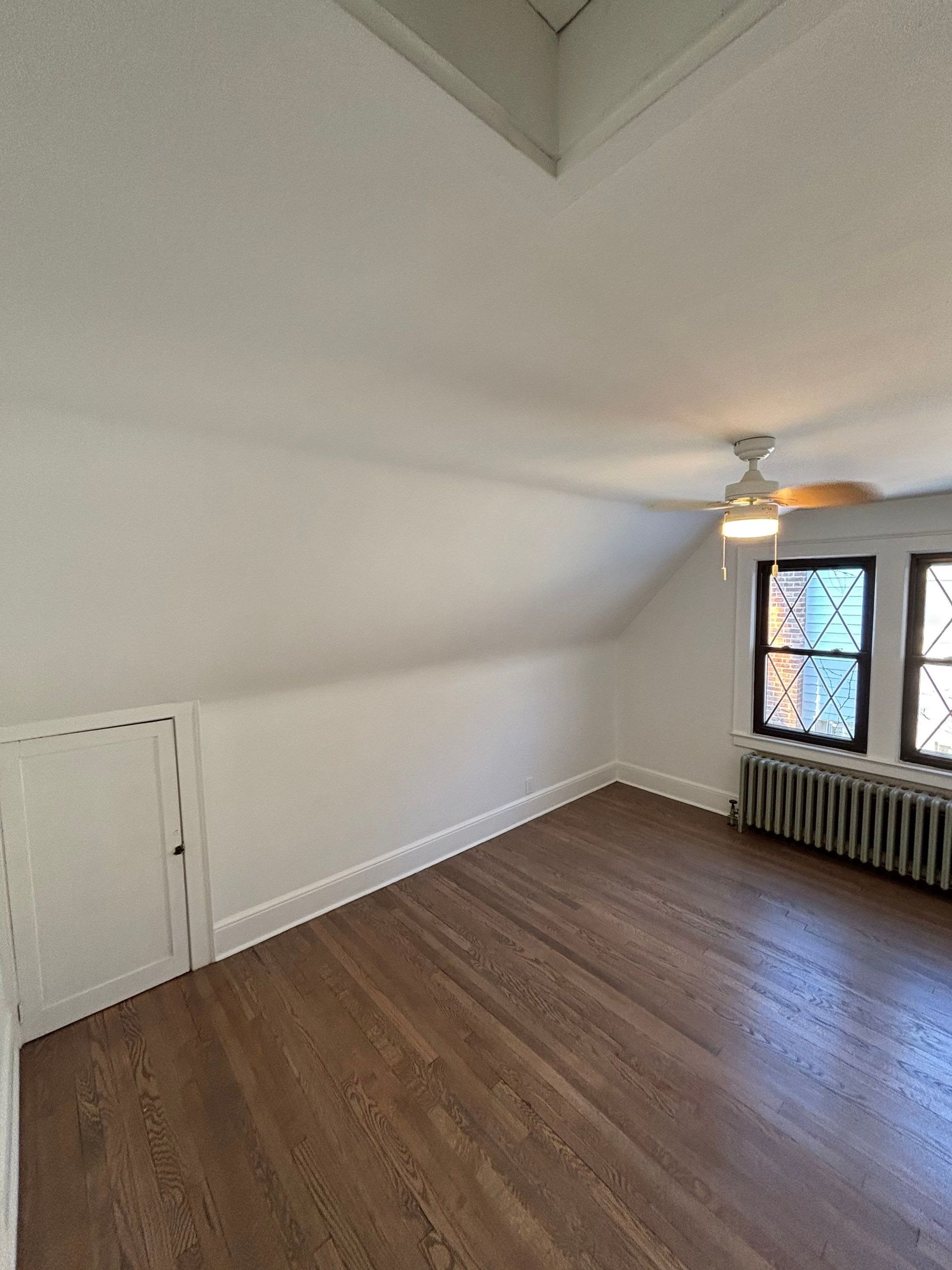 Empty room with hardwood floors, slanted ceiling, window with radiator.