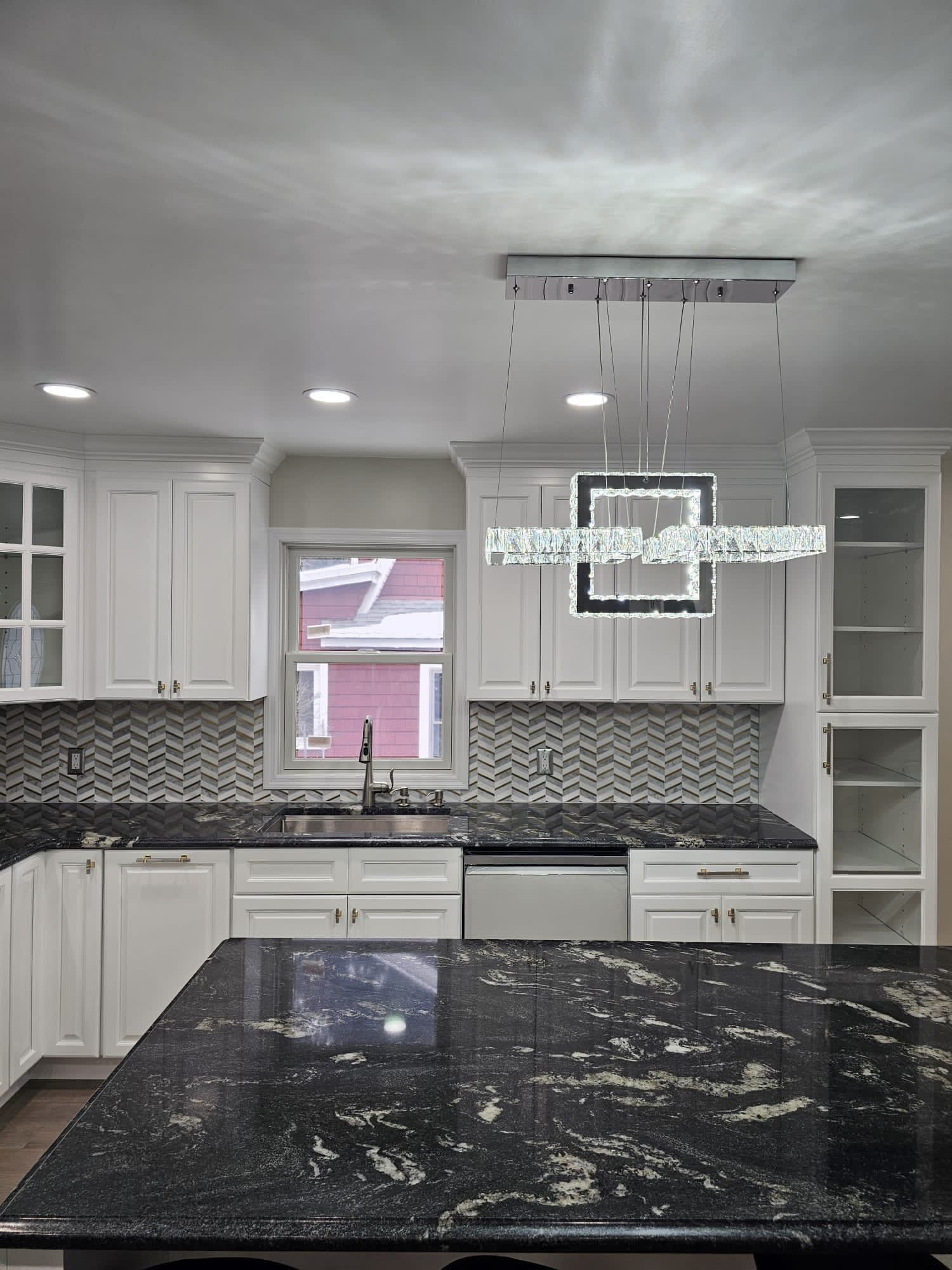White kitchen with black granite countertops, chevron backsplash, and modern crystal chandelier.