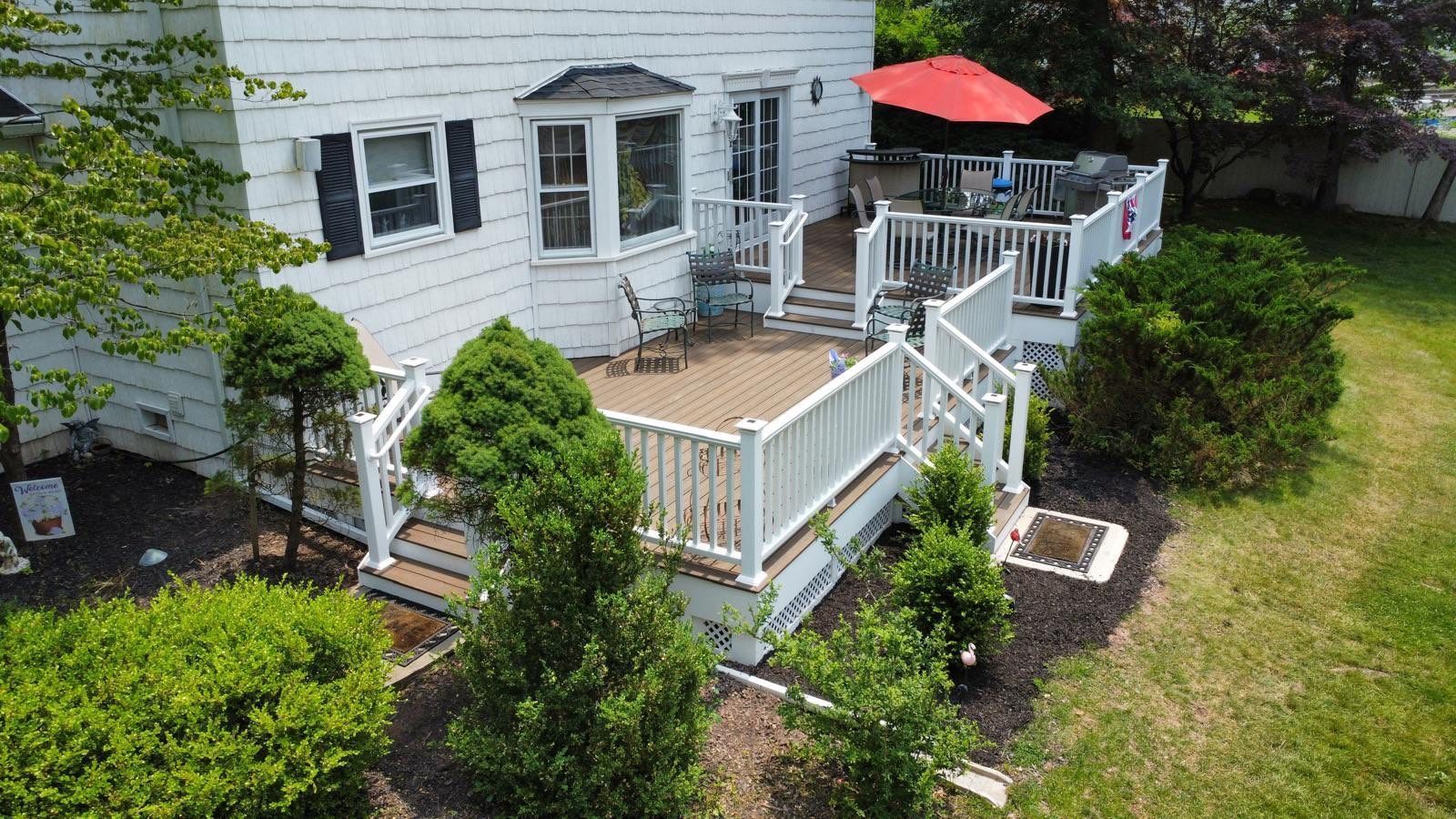 White house with wooden deck, small table, and red umbrella. Green grass surrounds.