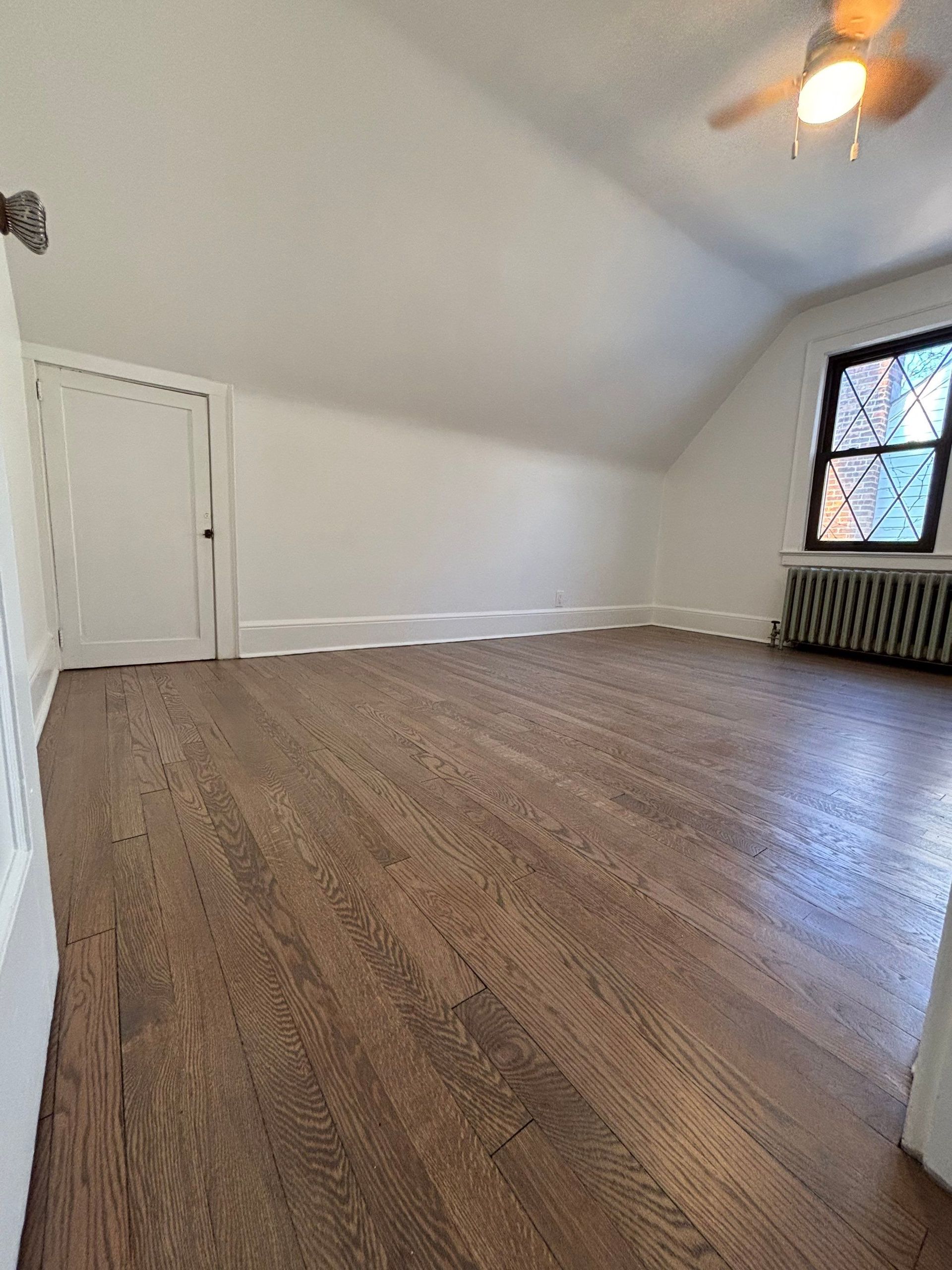 Empty attic room with dark wood floor, white walls, and a window.