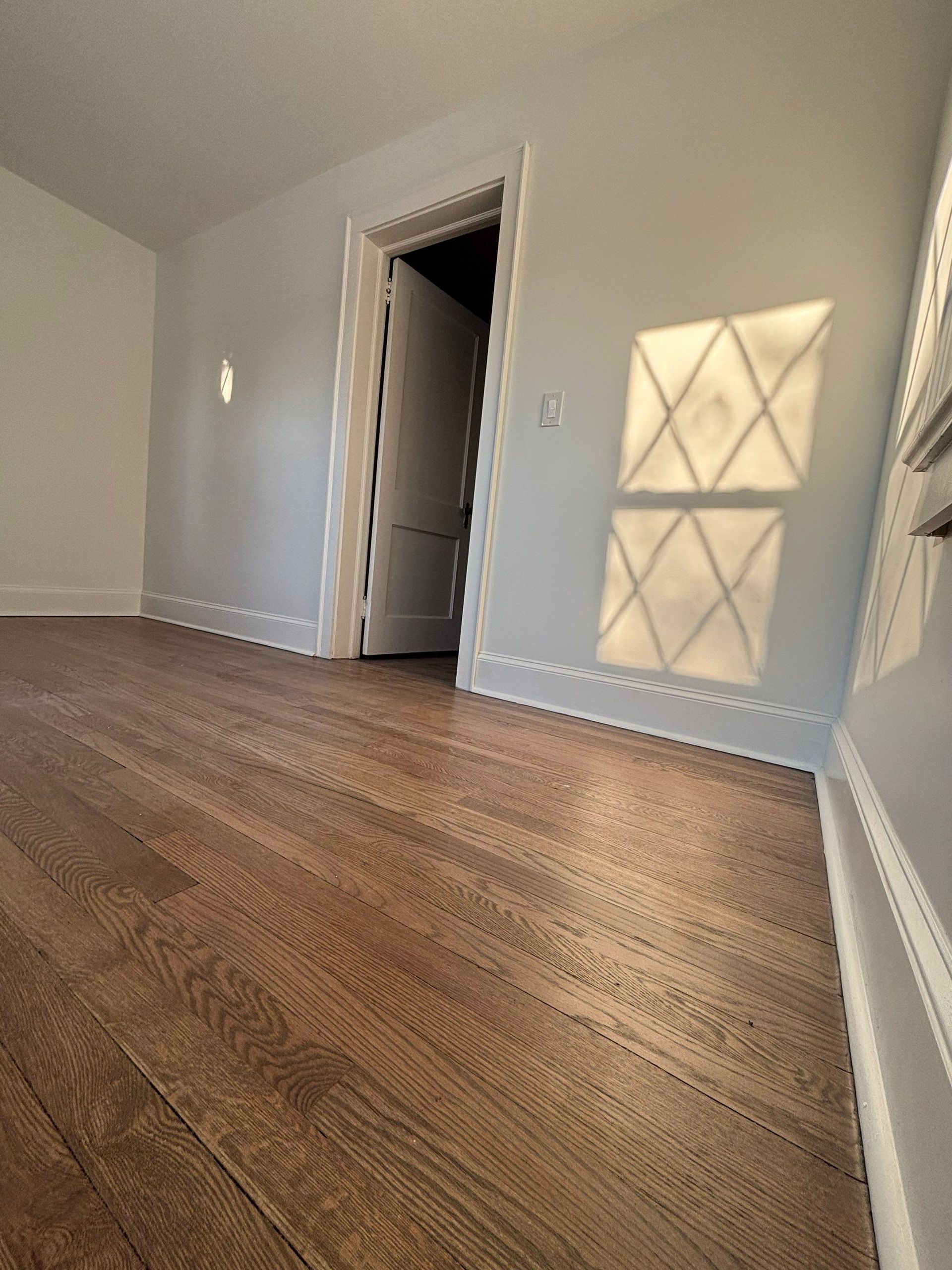 Empty room with hardwood floors, white walls, doorway, and sunlight casting a diamond pattern on wall.