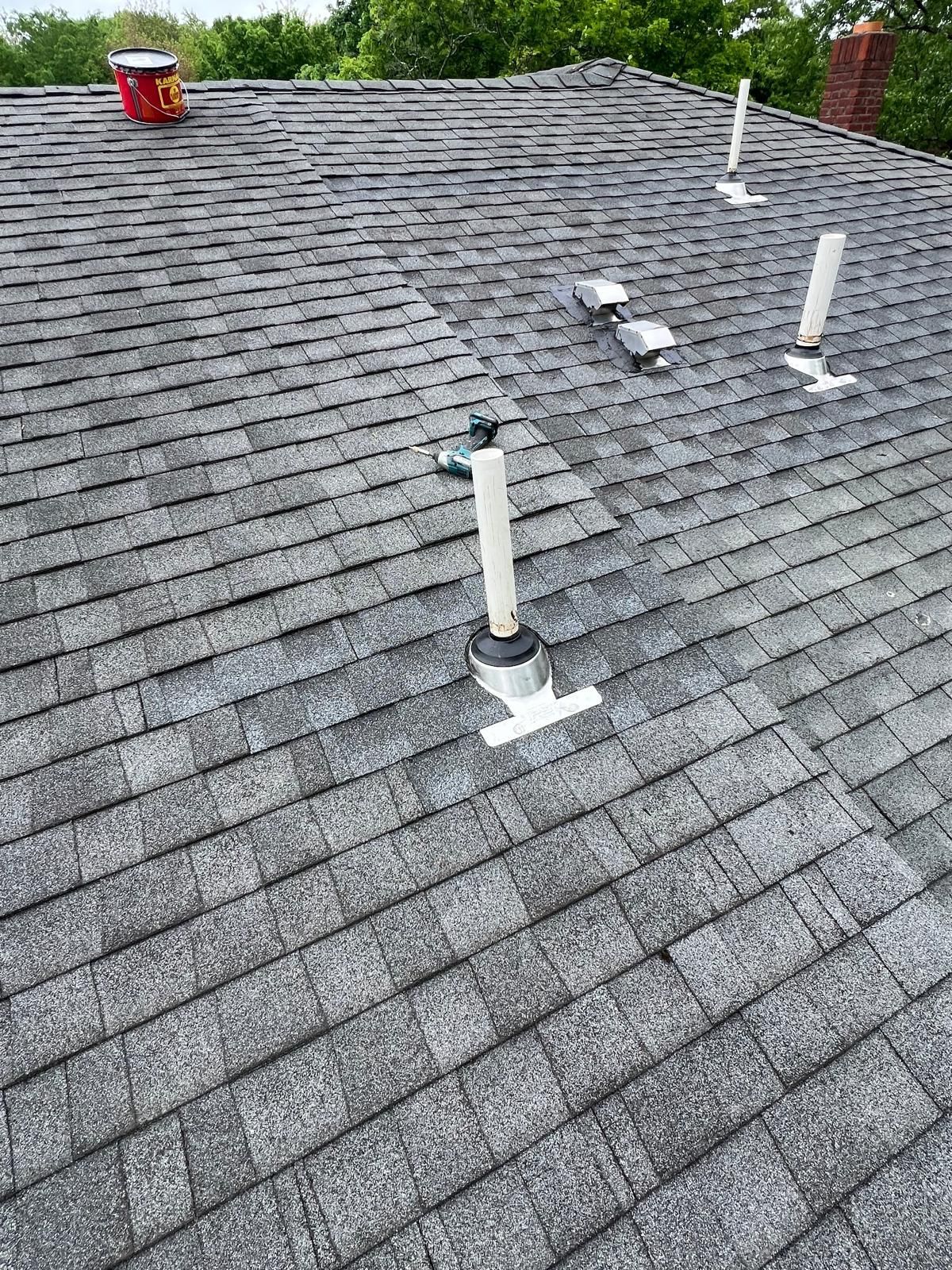 Gray shingled roof with multiple vent pipes and a bucket.
