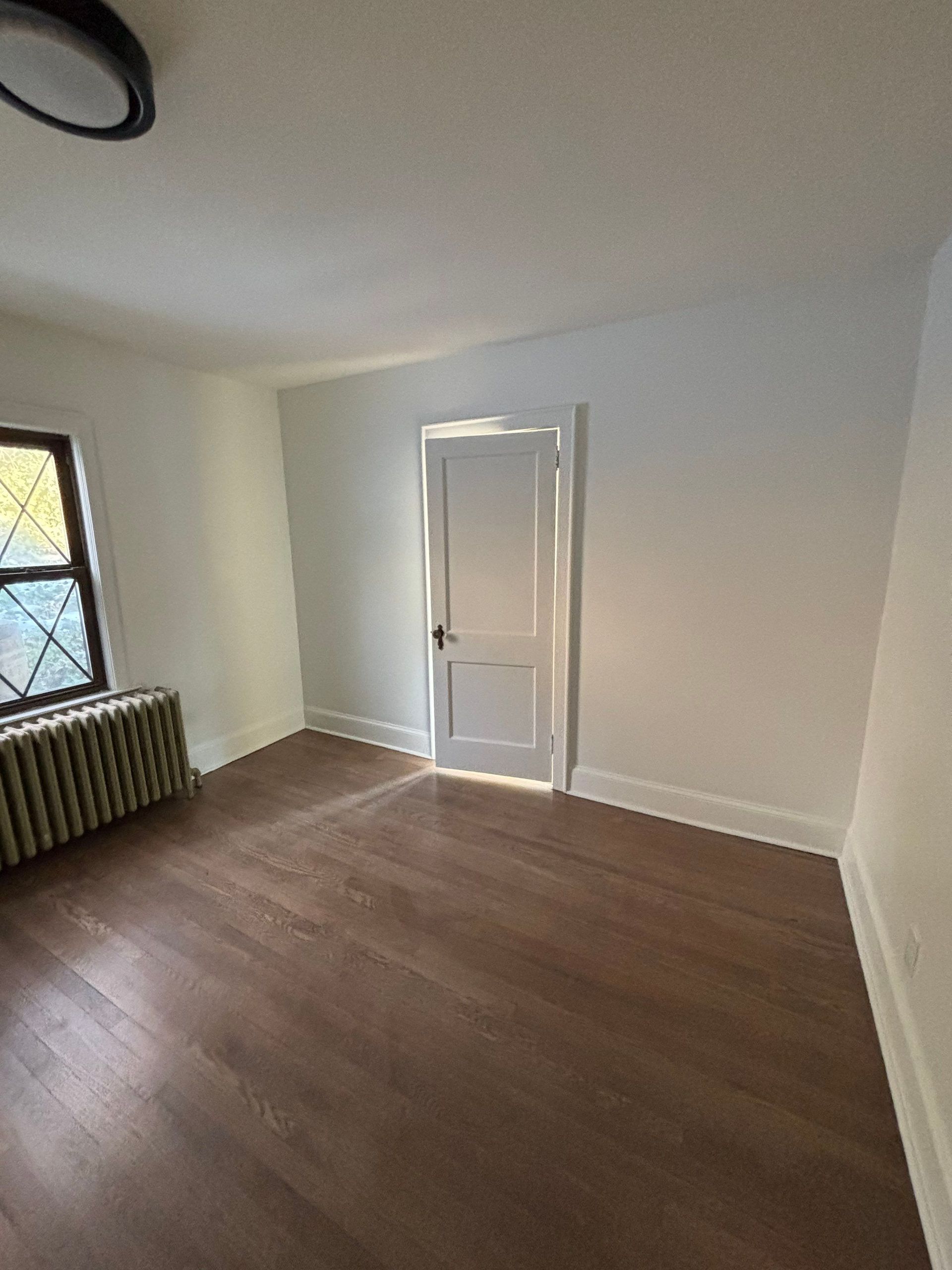Empty room with dark wood floor, white walls, radiator, and a door.