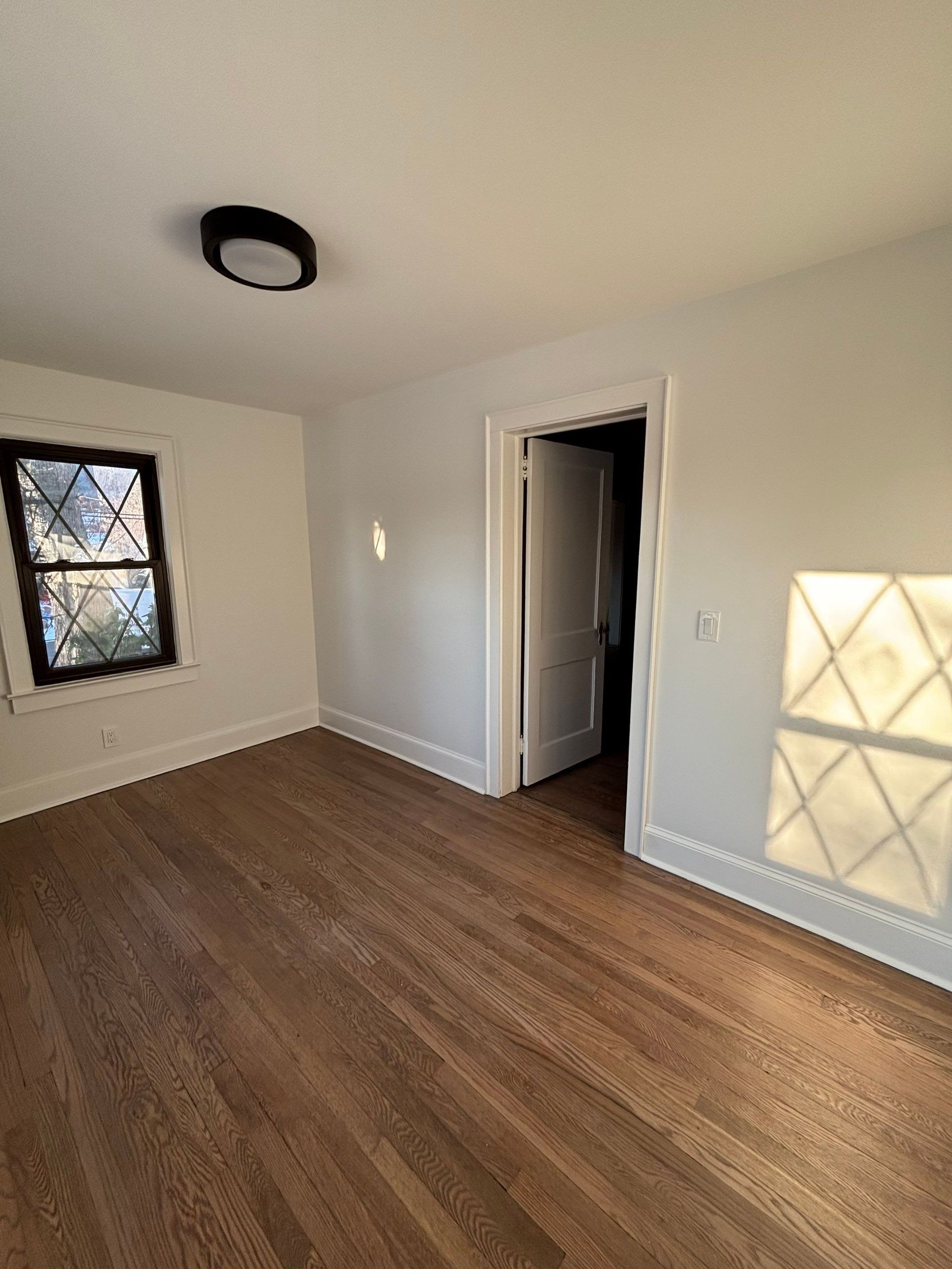 Empty room with hardwood floors, a closed door, and sunlight shining through a leaded glass window, illuminating the wall.