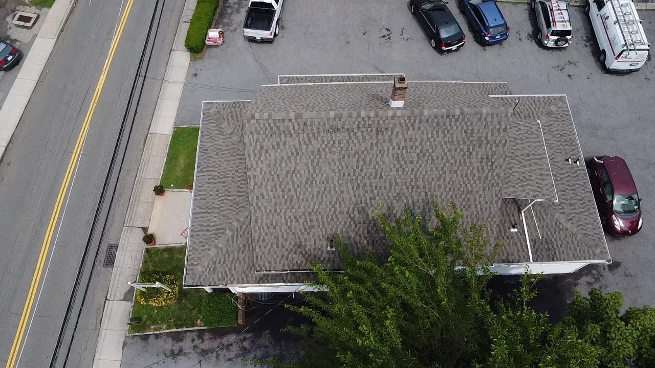 Aerial view of a building with a shingle roof and cars parked nearby. Street and landscaping visible.