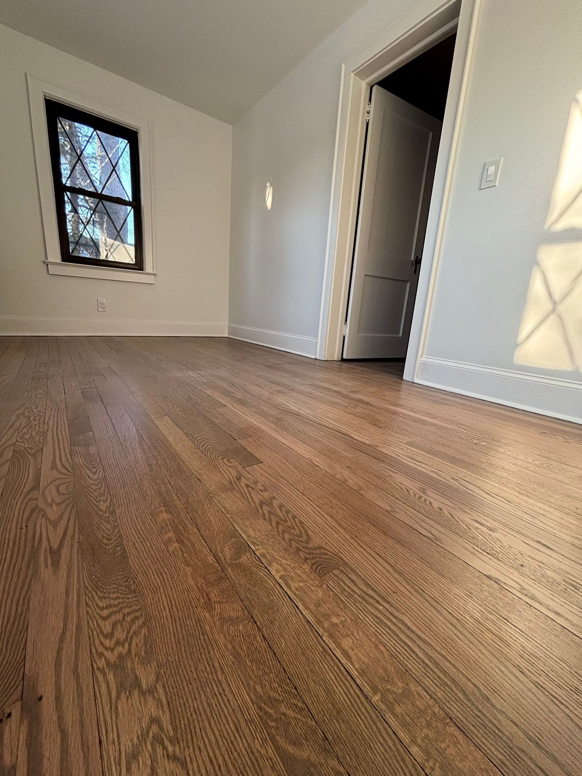 Empty room with hardwood floors, white walls, and a window. An open door is visible.