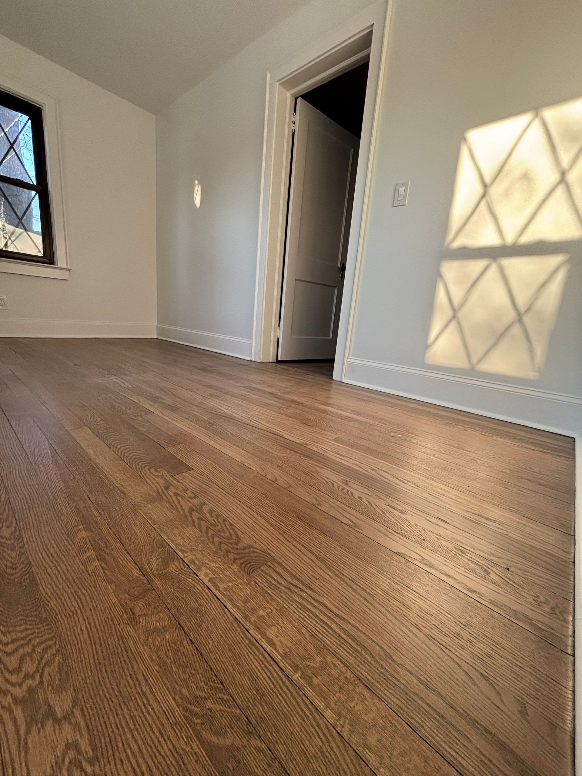 Hardwood floor in room with white walls, sunlight casting a diamond pattern shadow.