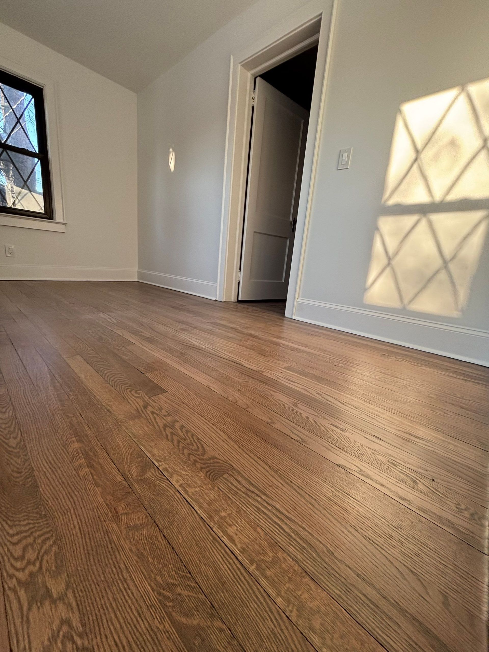 Hardwood floor in an empty room, white walls, sunlight through a window casting shadows.