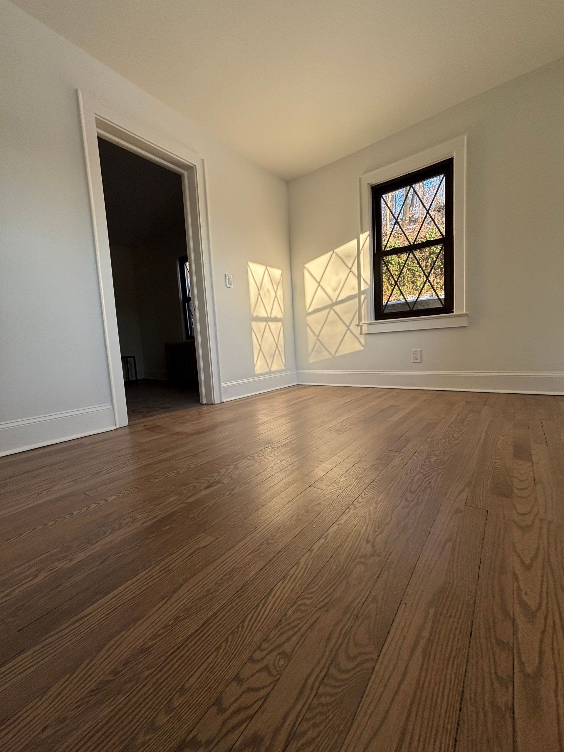 Empty room with hardwood floors, white walls, doorway, and window casting light.
