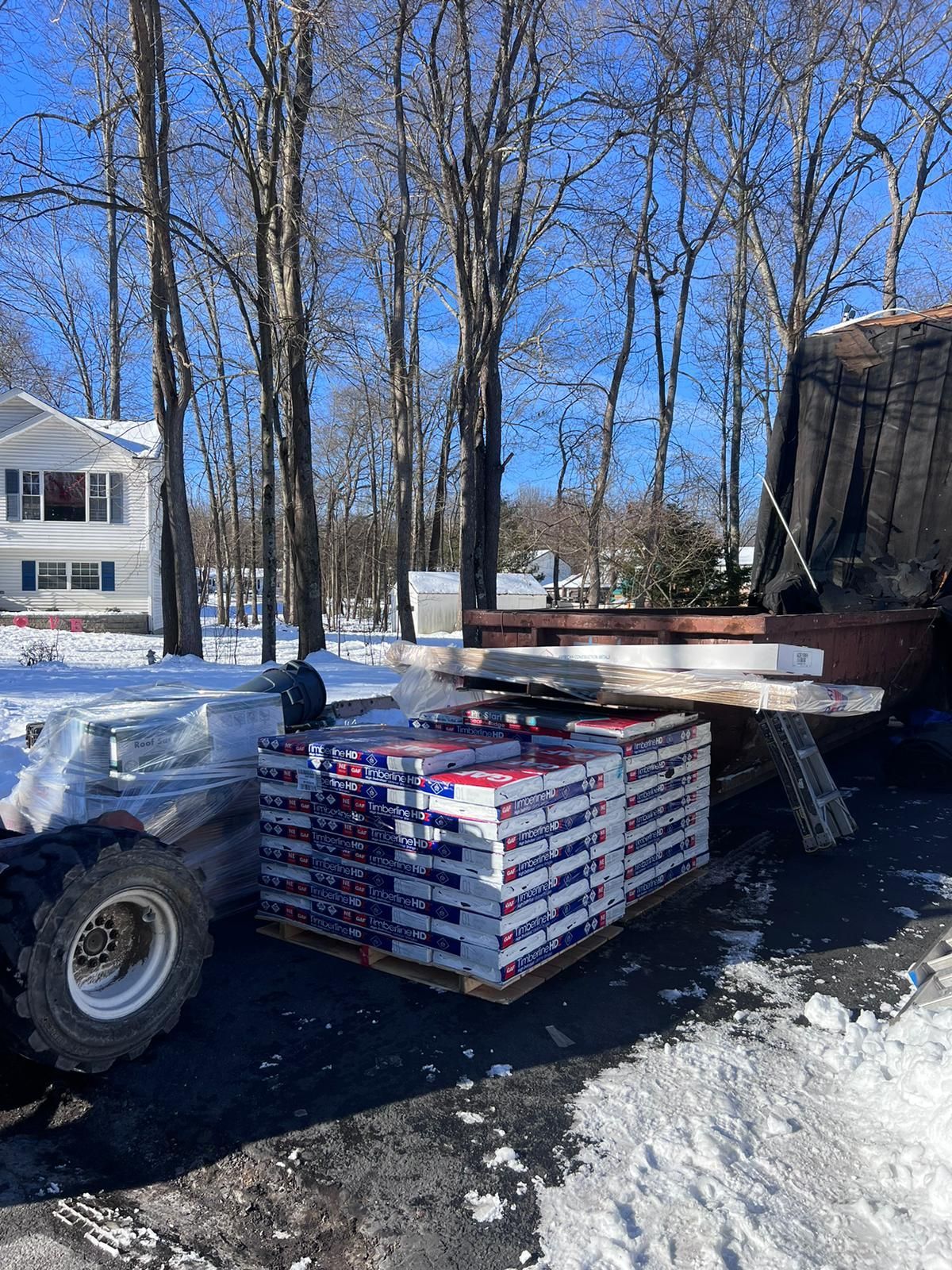 Pallet of boxes on snow, forklift, house, and trees under a clear blue sky.
