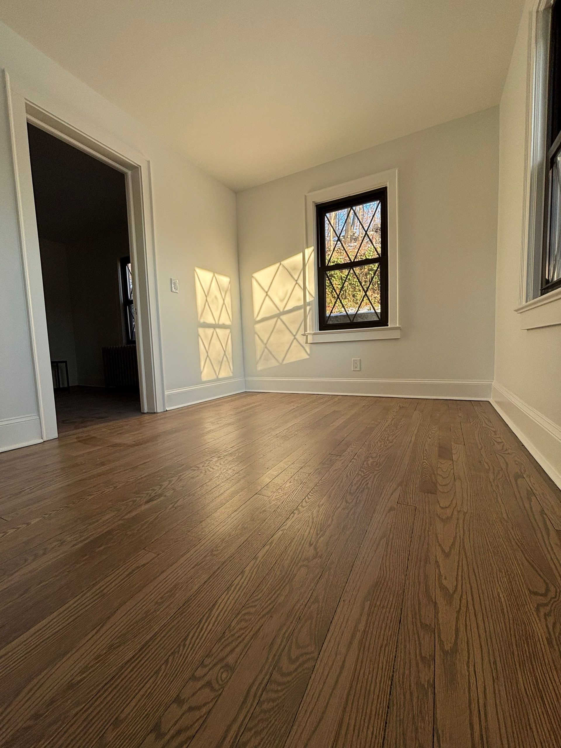 Empty room with hardwood floors, a window, and a door opening. Sunlight creates geometric shadows.