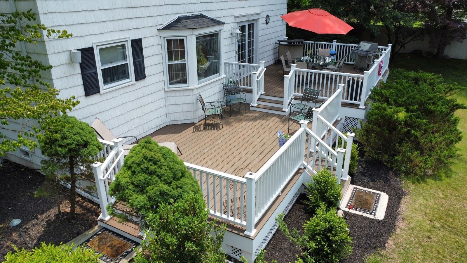 White house with multi-level deck, white railings, brown decking. Red umbrella, chairs, shrubbery. Sunny day.