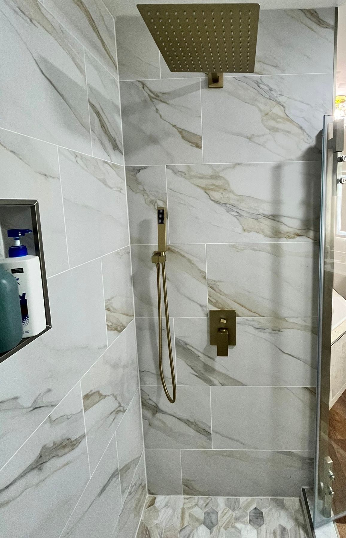 Shower with gold fixtures and marble-look tiles. Includes a built-in shelf and glass door.