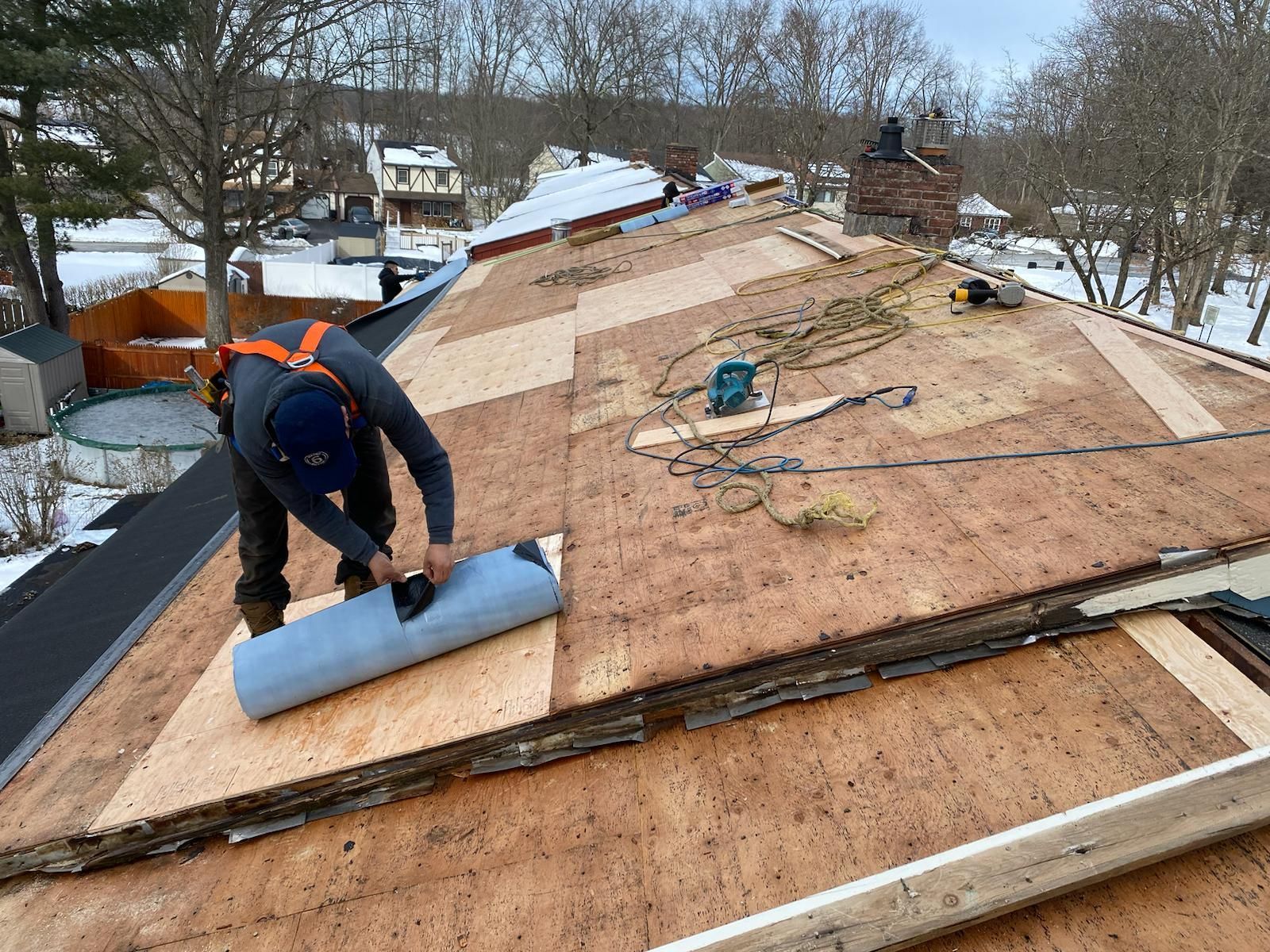 Roofer on a partially-demolished roof, installing roofing underlayment. Snowy yard in the background.