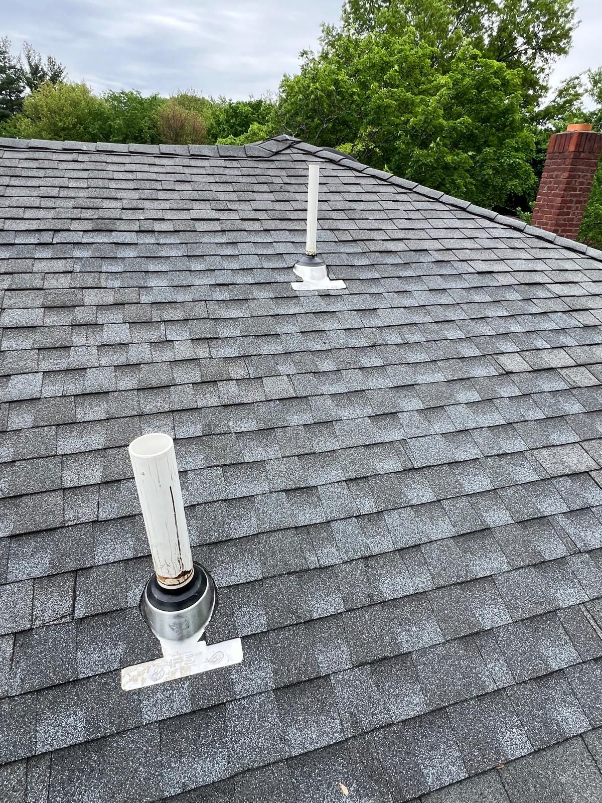 Gray shingle roof with two white PVC vent pipes and a red brick chimney in the background.