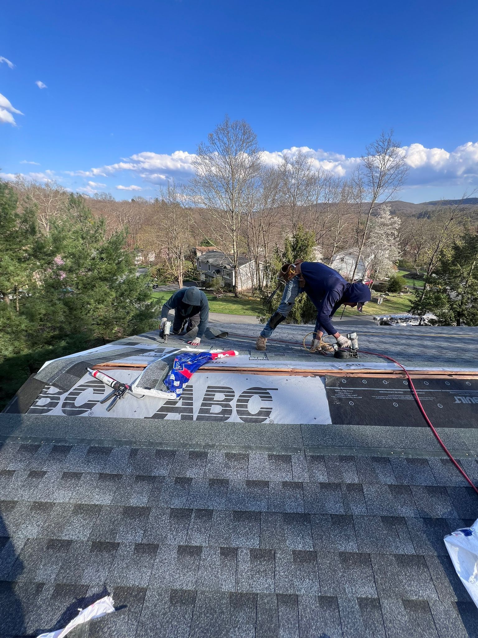 Two roofers installing shingles on a house roof under a sunny, blue sky.