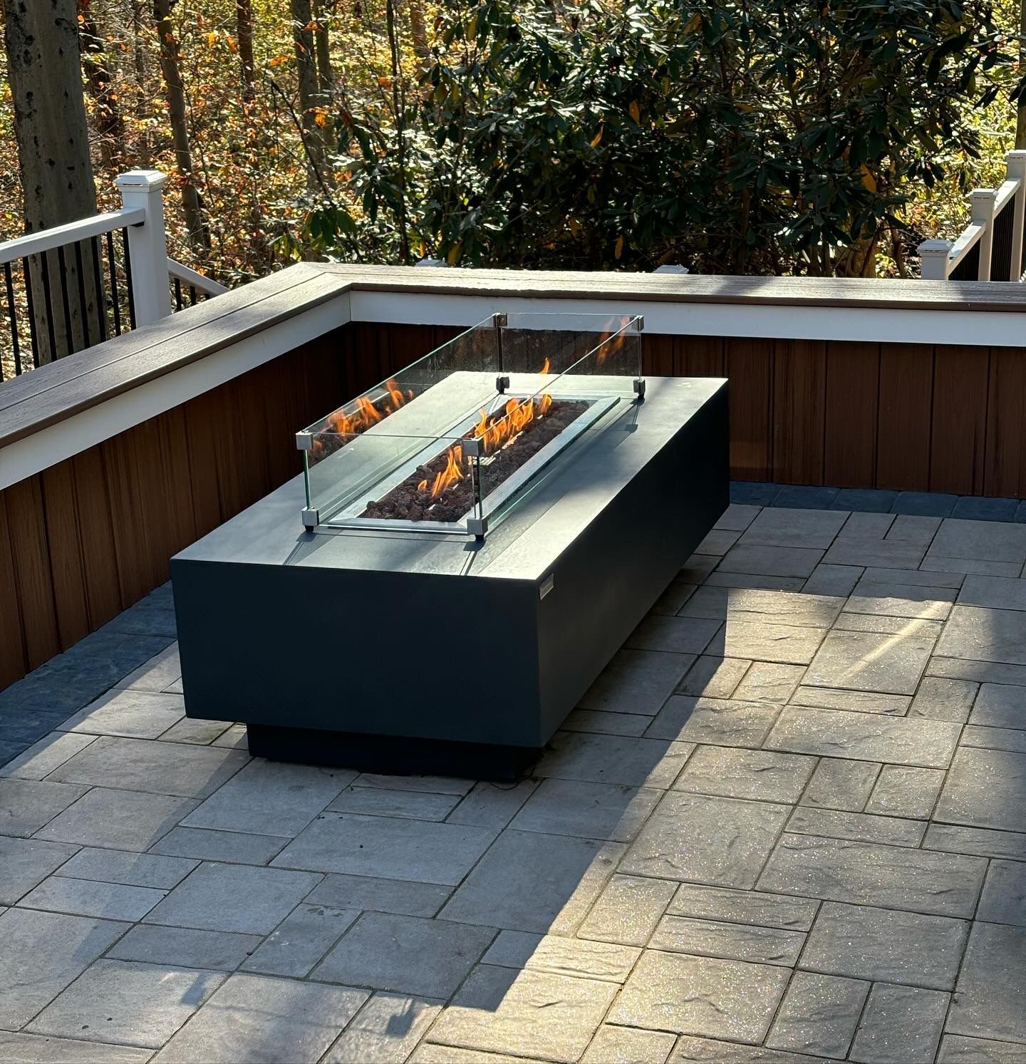 Rectangular fire pit with glass wind guard on a patio. Deck and trees in the background.