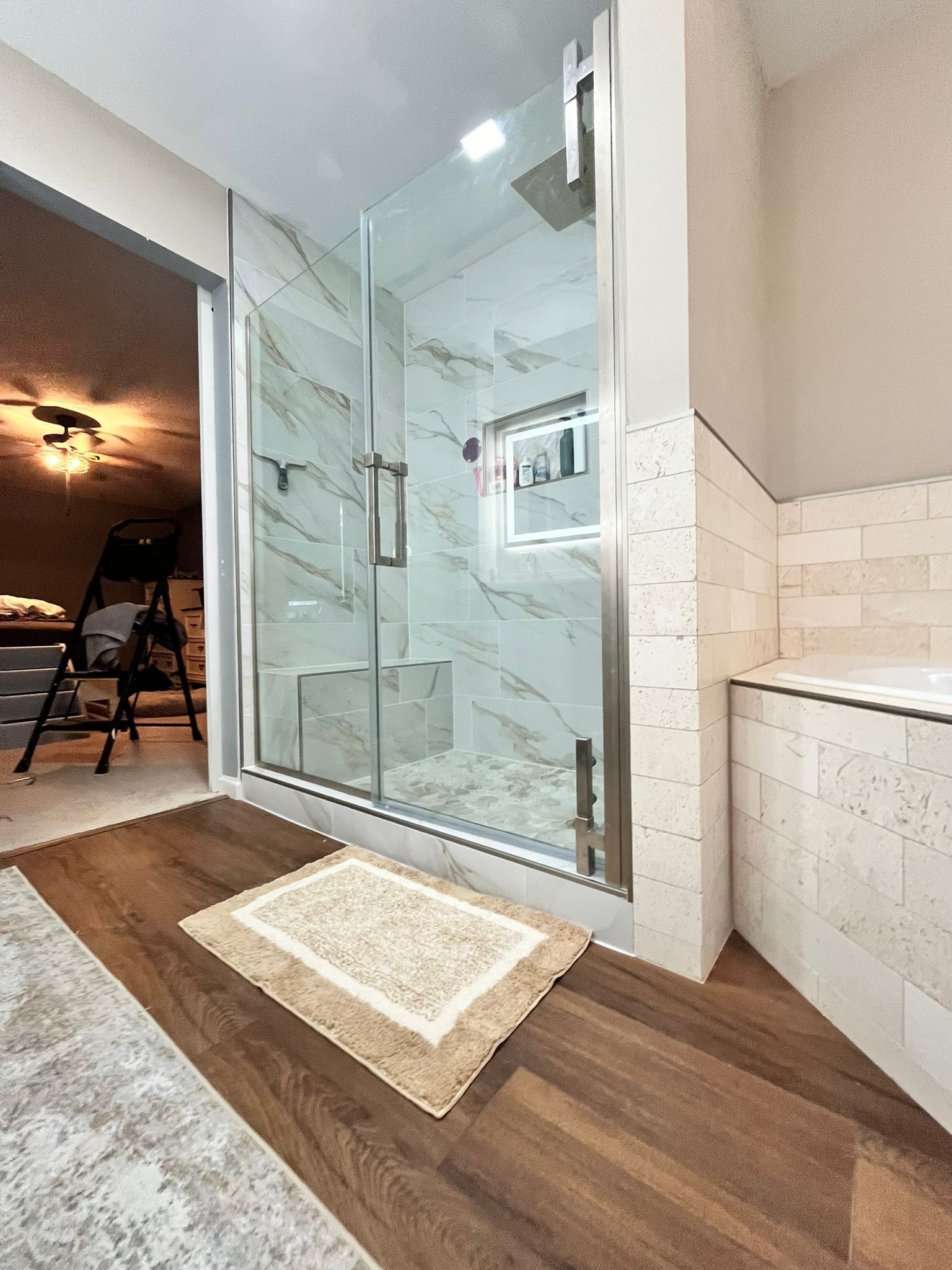 Bathroom with glass shower, marble tile, wood floor, and beige rug.