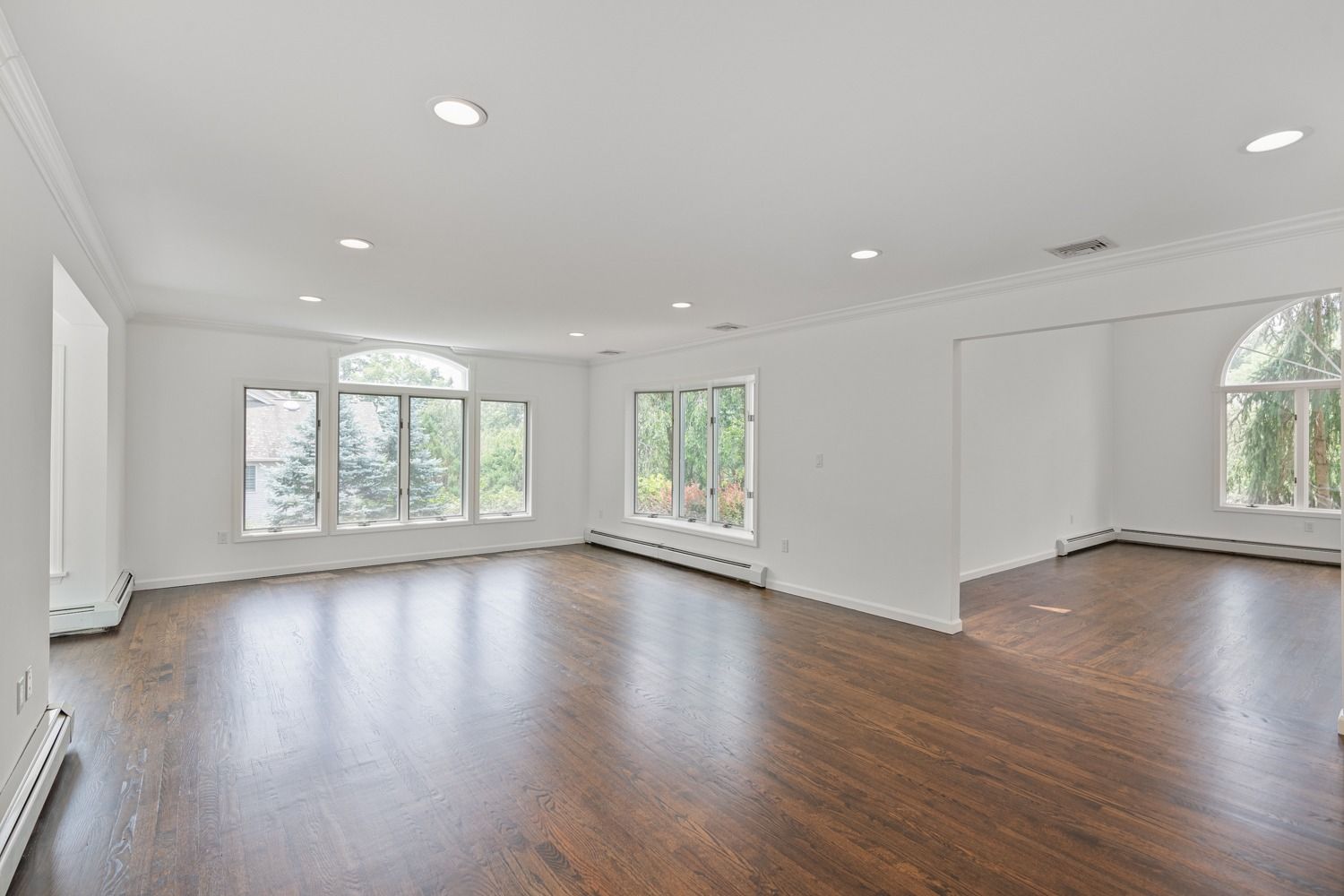 Spacious, empty room with dark wood floor, white walls and trim, and large windows overlooking trees.