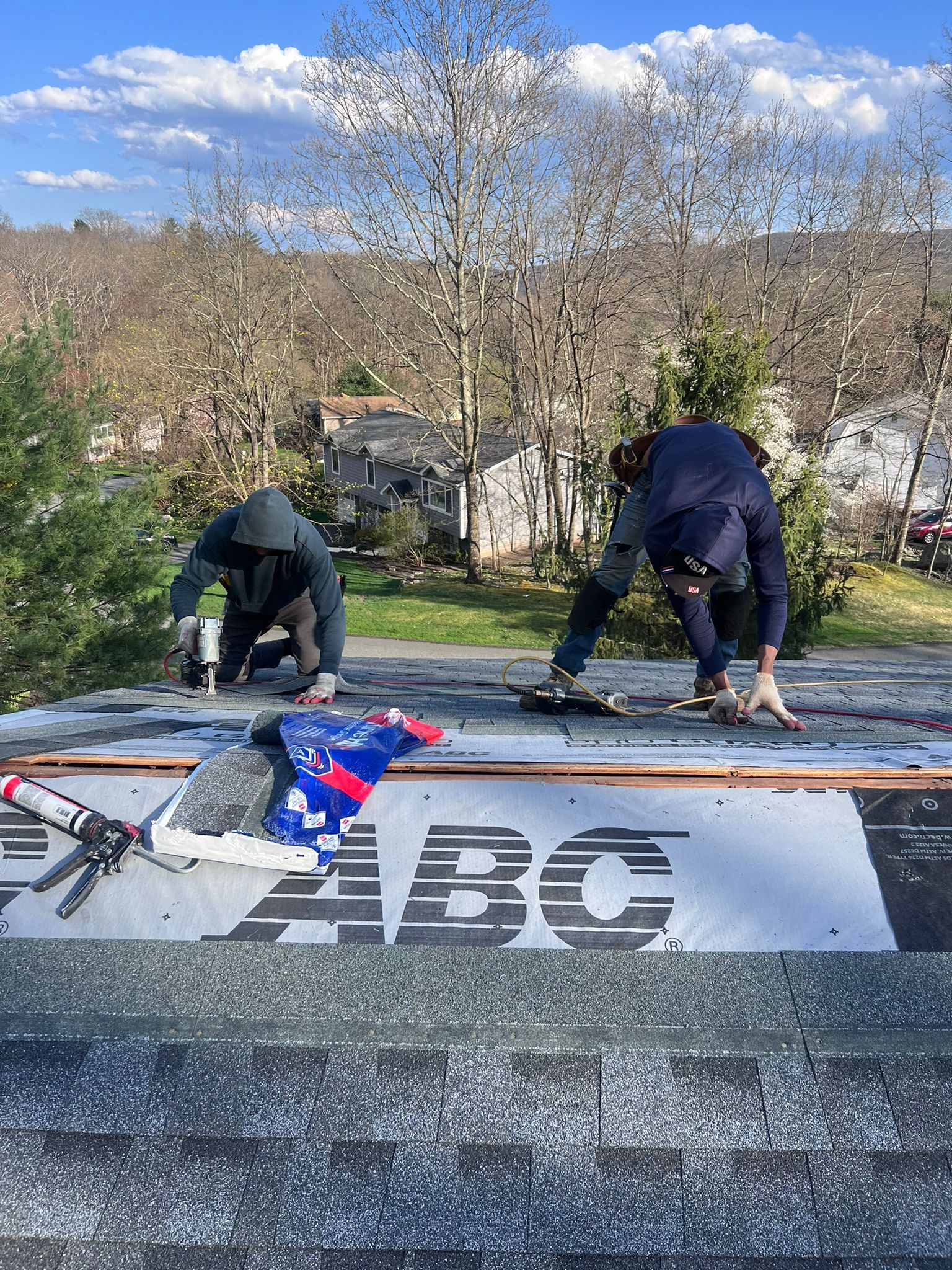 Two people installing shingles on a roof under a blue sky, ABC logo on underlayment.