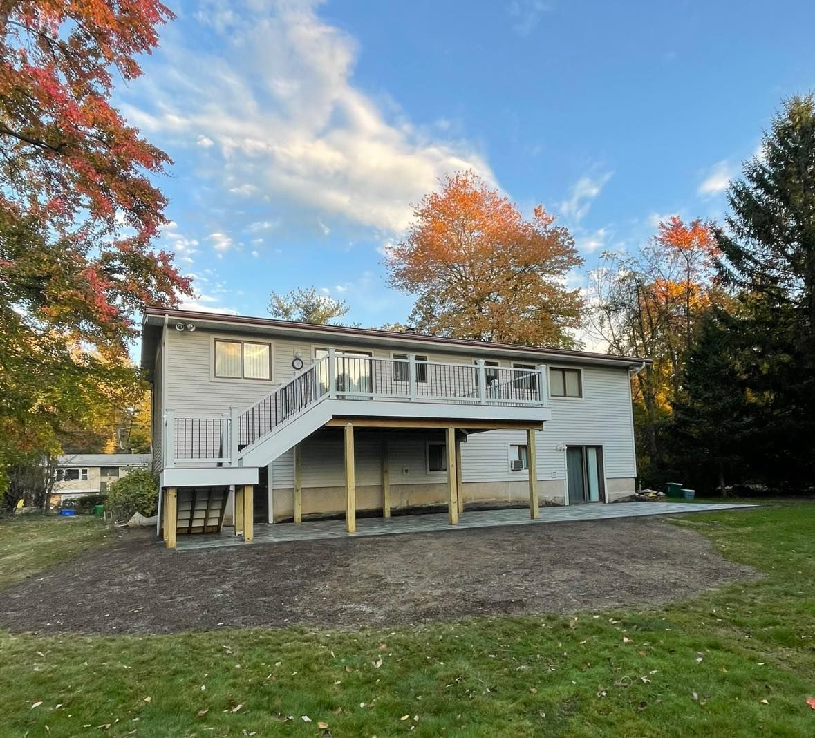 Back of a two-story house with a wooden deck and patio. Autumn trees surround the house under a blue sky.