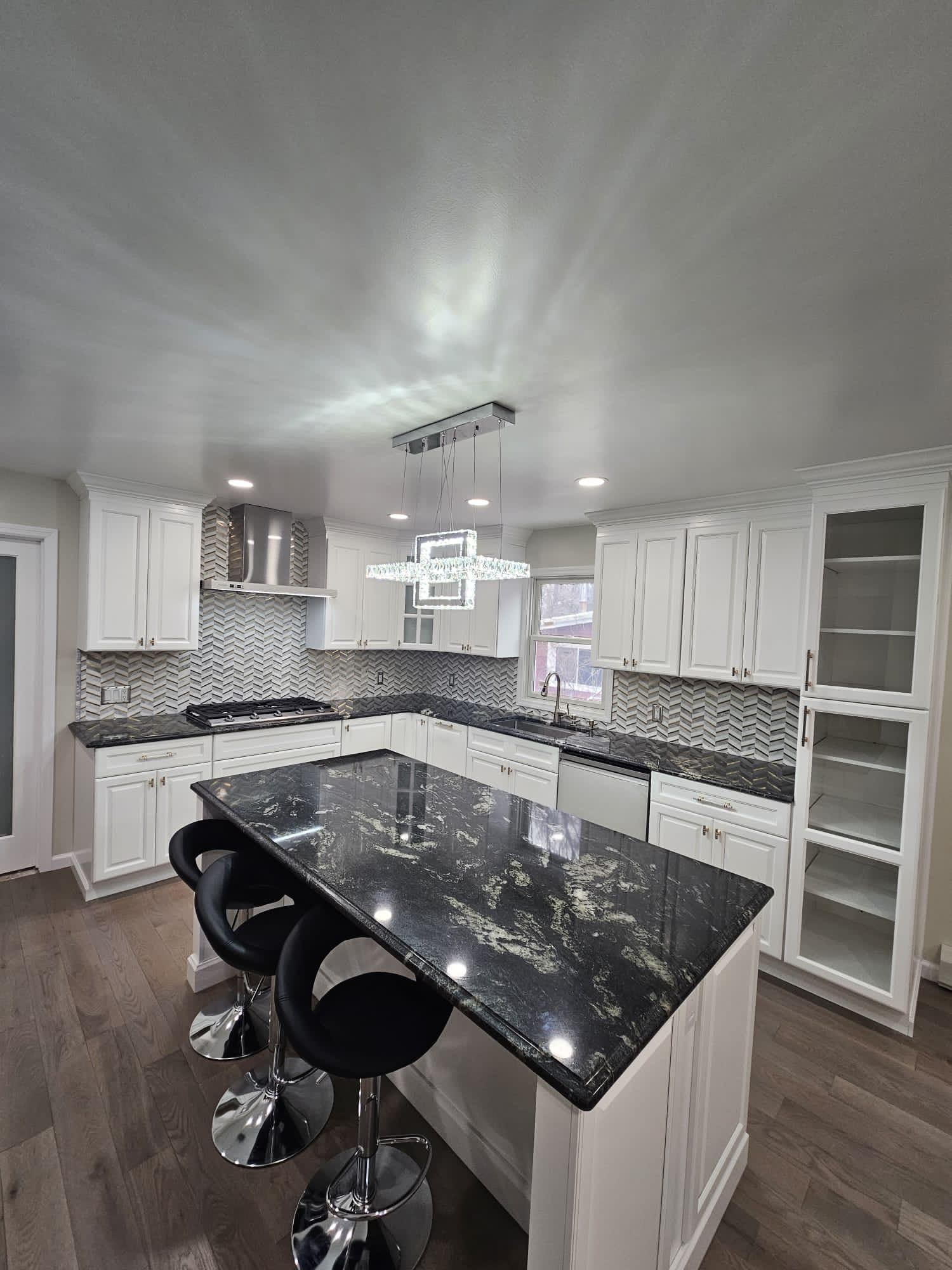 Modern white kitchen with black granite island and bar stools, stainless steel appliances, and wood flooring.