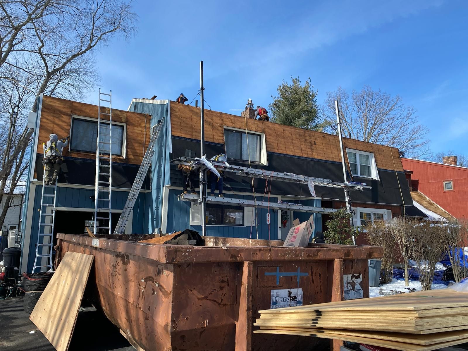 Workers on a blue house roof, removing shingles. A large dumpster sits below, collecting debris.