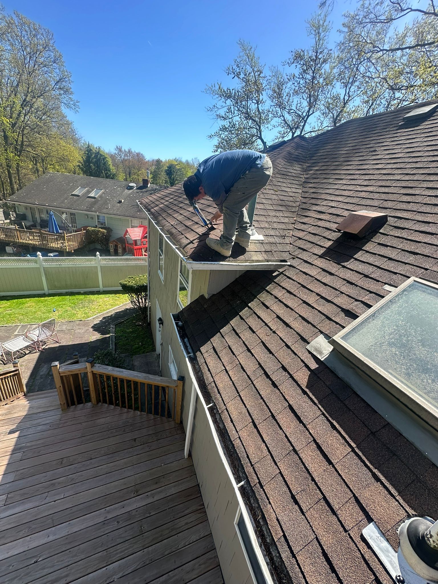 Person on a roof repairs shingles on a sunny day. House with deck and trees visible.