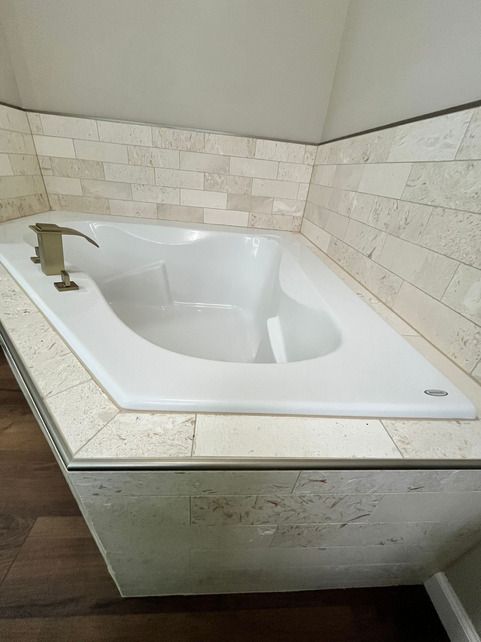 White bathtub surrounded by light beige tile, set into a corner. Gold faucet.
