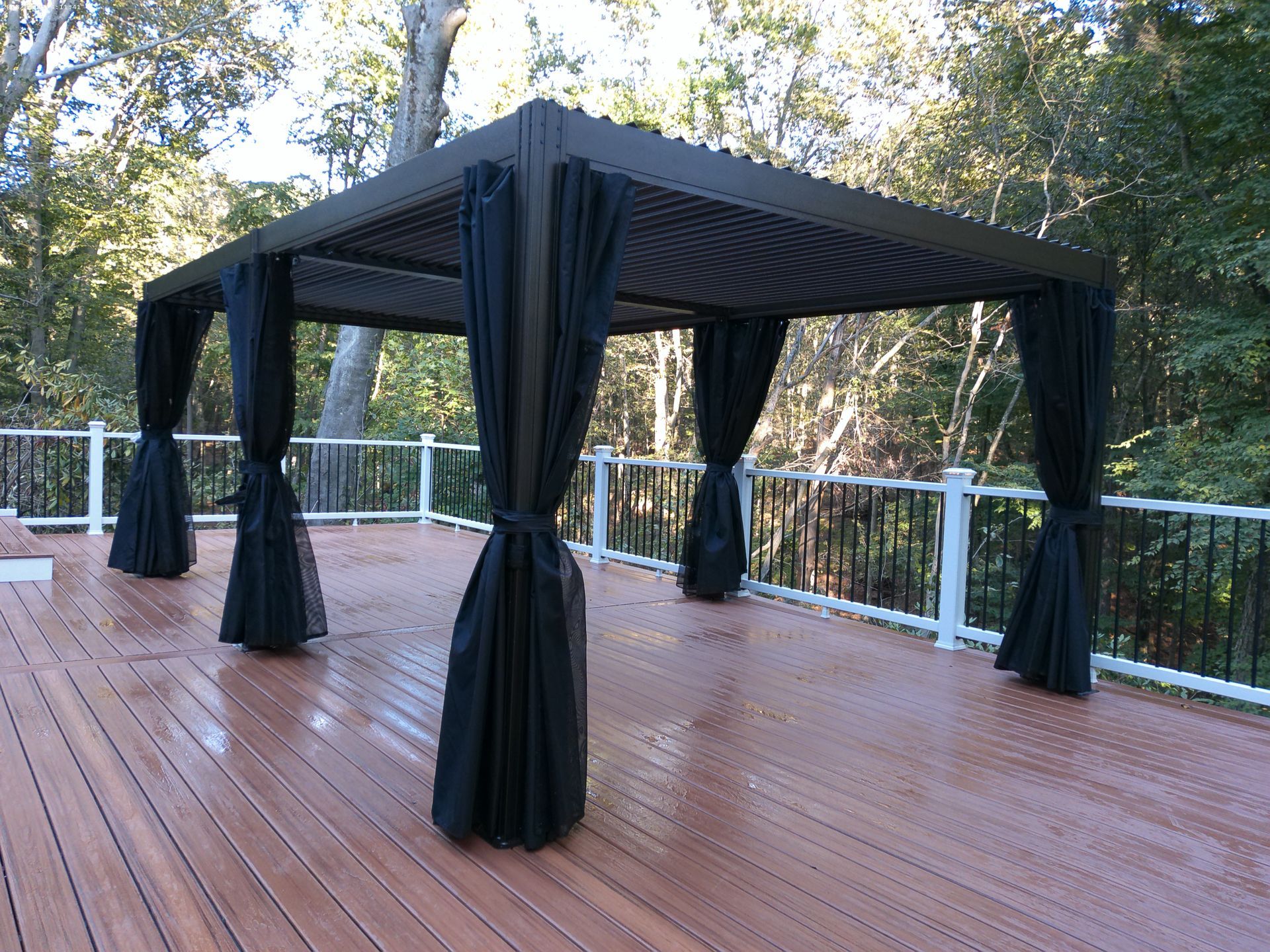 Black pergola with black curtains on a wooden deck, set against a backdrop of trees.
