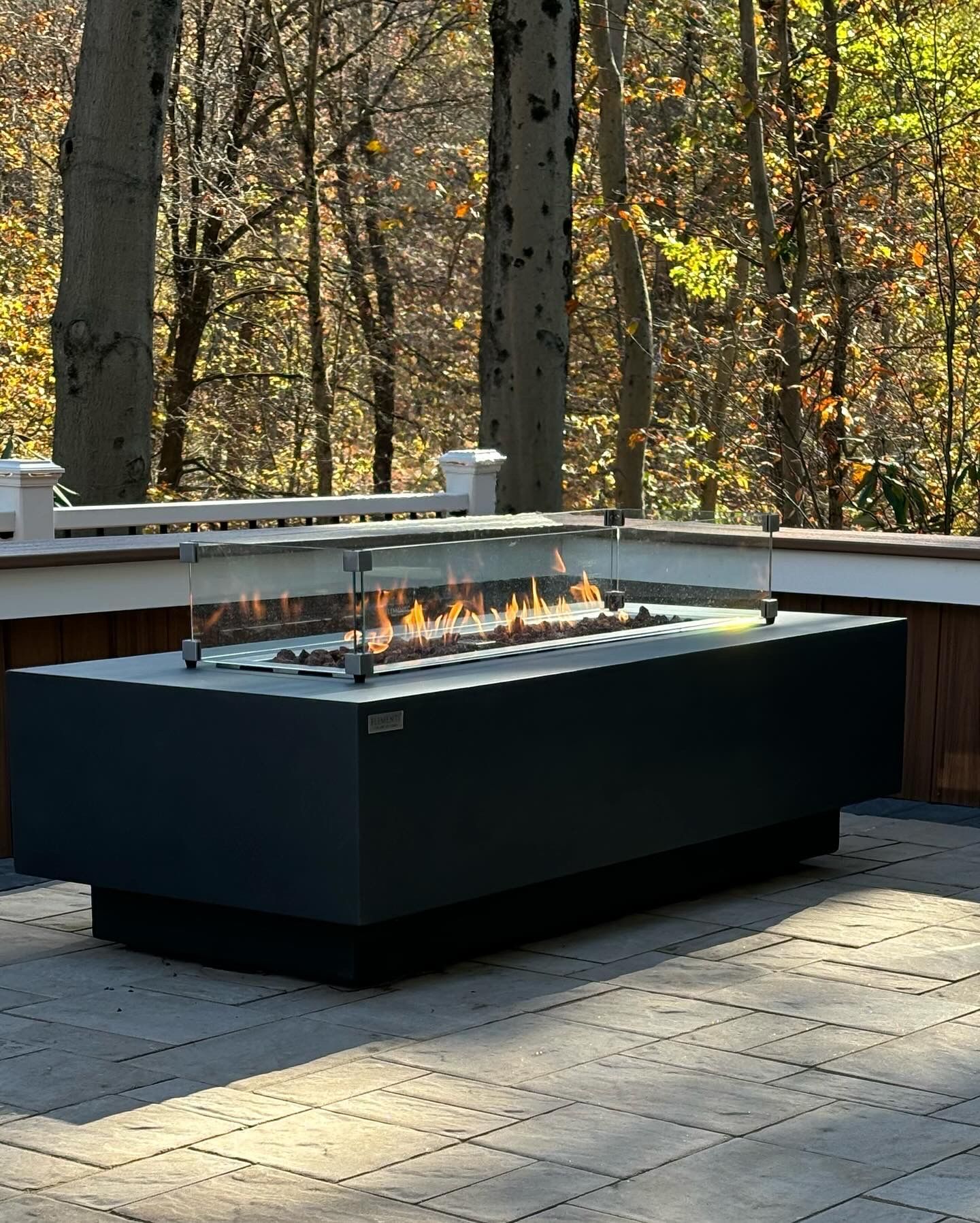 Rectangular outdoor fire pit with flames, on a stone patio, next to a deck, and trees in the background.