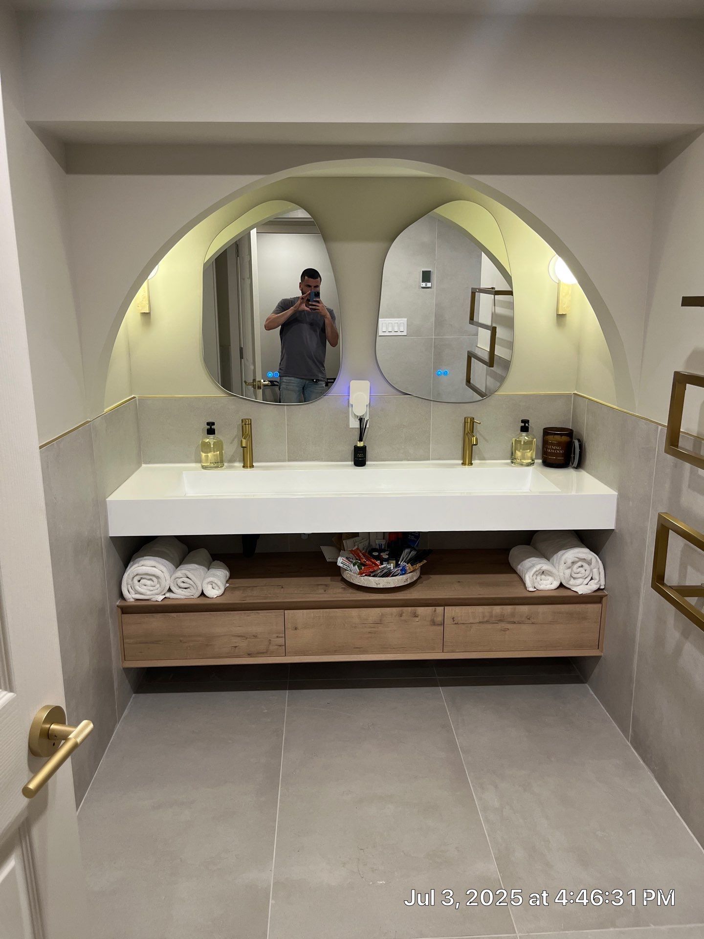 Bathroom with double sink, arched mirrors, gold fixtures, light wood vanity, and a person taking a photo in the mirror.