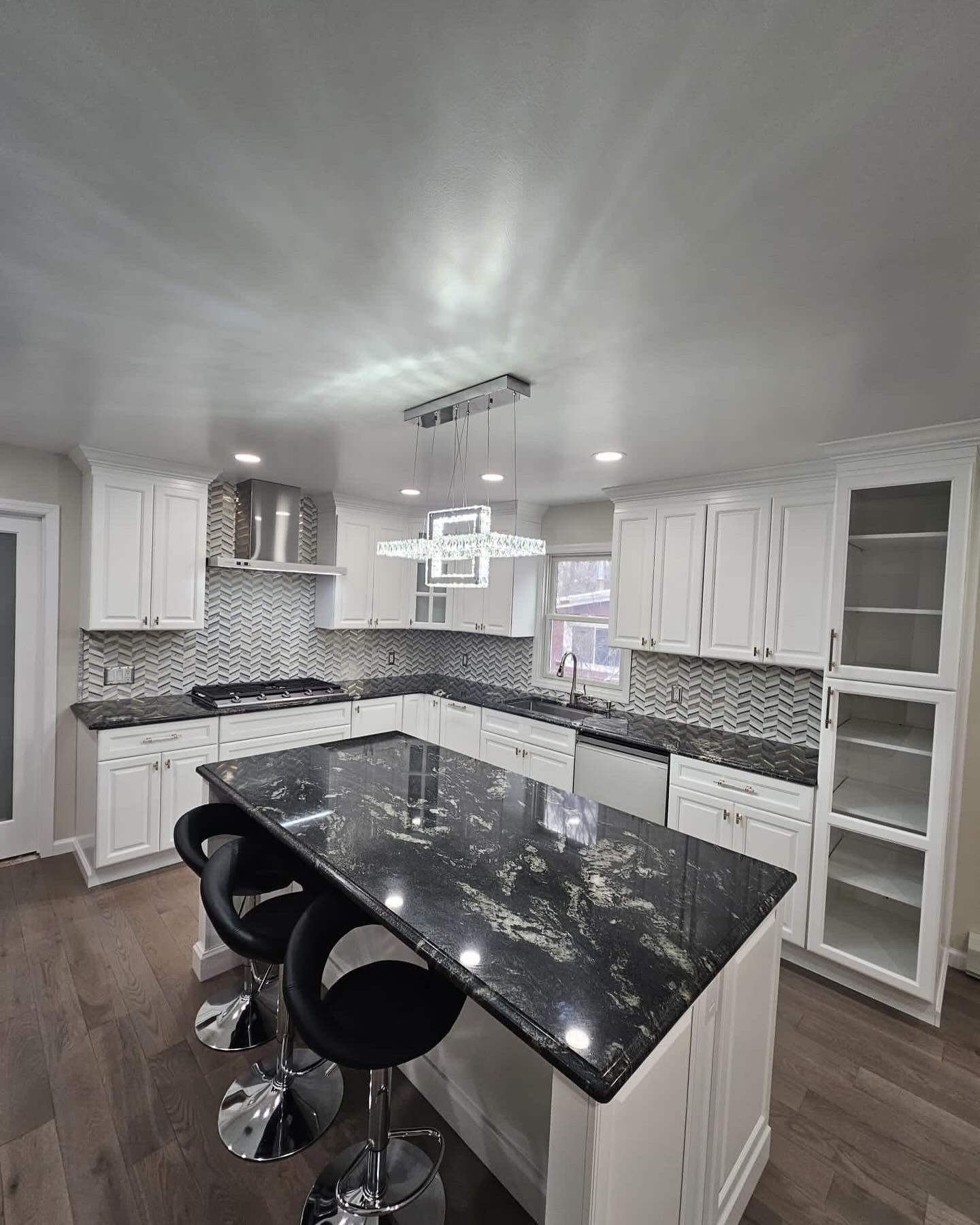 Modern white kitchen with a granite island, black bar stools, and overhead lighting.